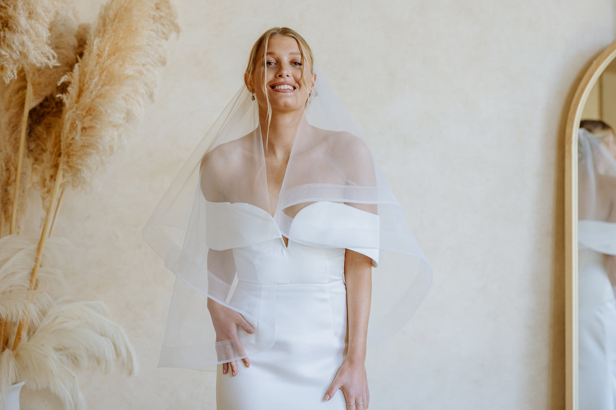 A woman wearing a white off-the-shoulder wedding dress and a sheer white wedding veil, standing indoors against a warm beige wall with dried pampas grass on the left and part of a mirror on the right.