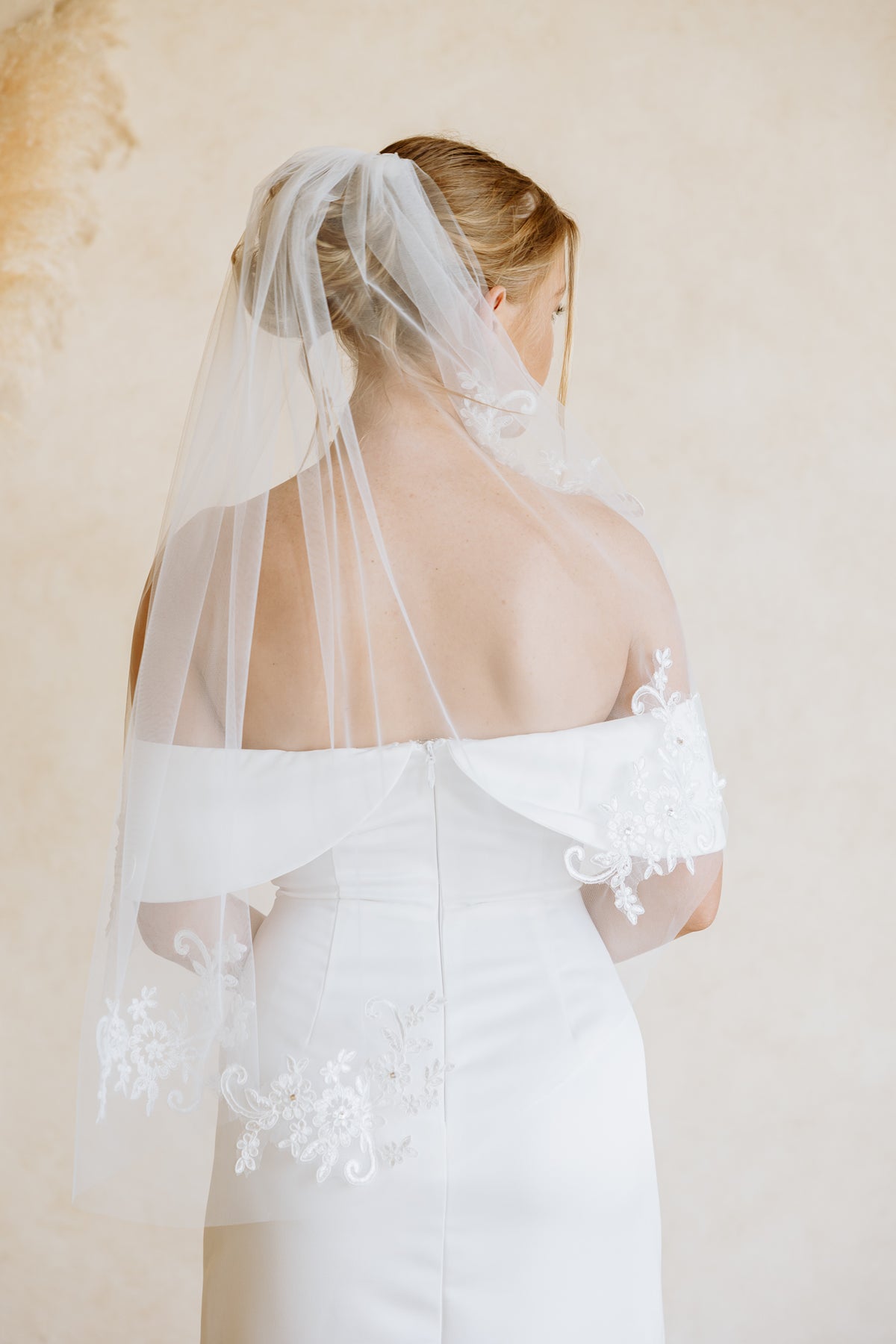 A bride viewed from behind wearing a white off-the-shoulder wedding dress and a sheer white veil with lace applique draped over her shoulders, set against a warm beige textured background.