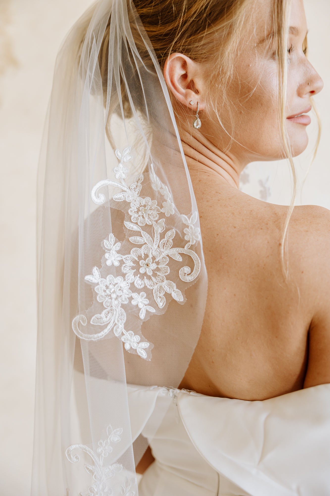 A bride viewed from behind wearing a white off-the-shoulder wedding dress and a sheer white veil with lace applique draped over her shoulders, set against a warm beige textured background.