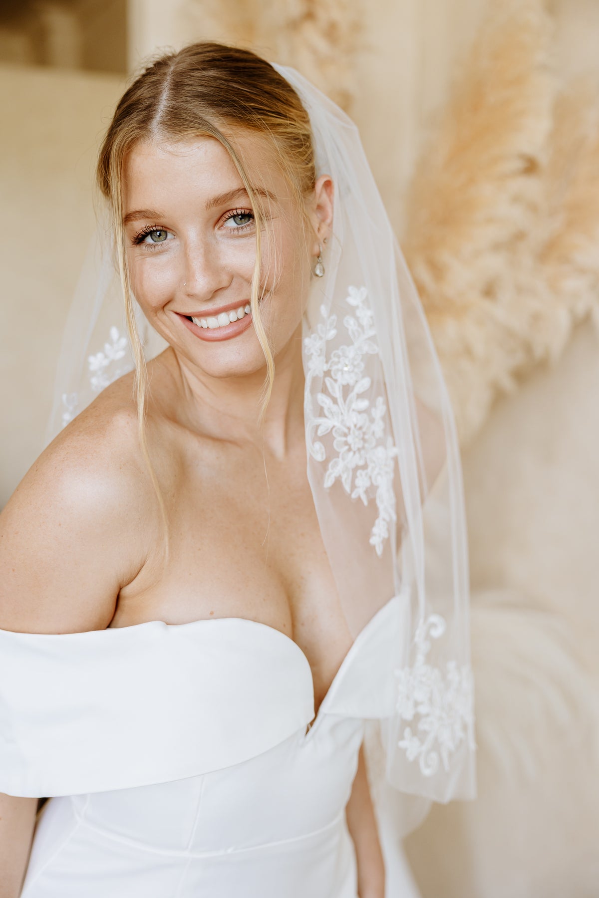 A smiling bride wearing a strapless white wedding dress and a sheer white lace-trimmed veil, photographed in a softly lit indoor setting with warm neutral decor.