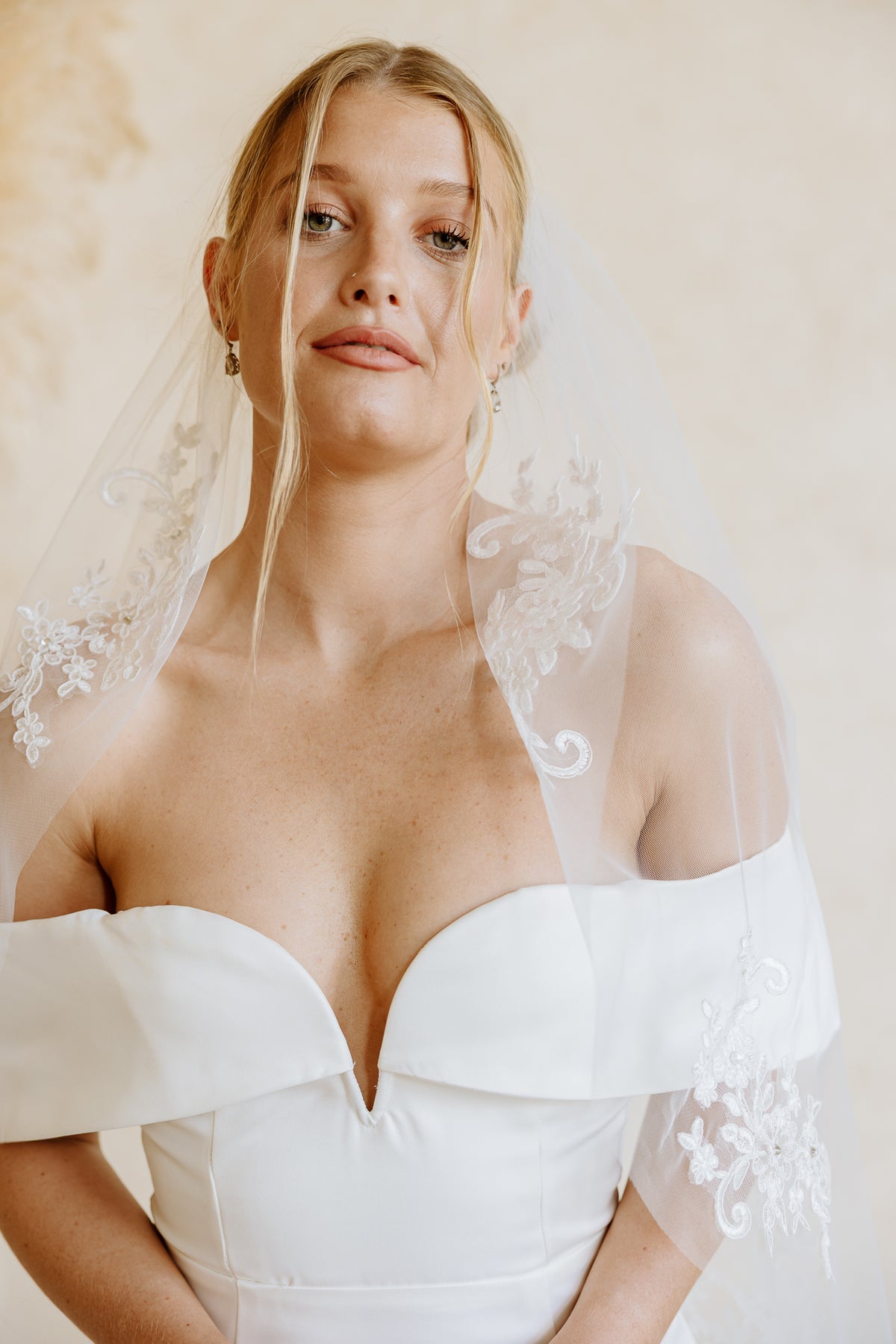A woman wearing a white off-the-shoulder wedding dress and a sheer white veil with floral lace trim, photographed against a soft beige background.