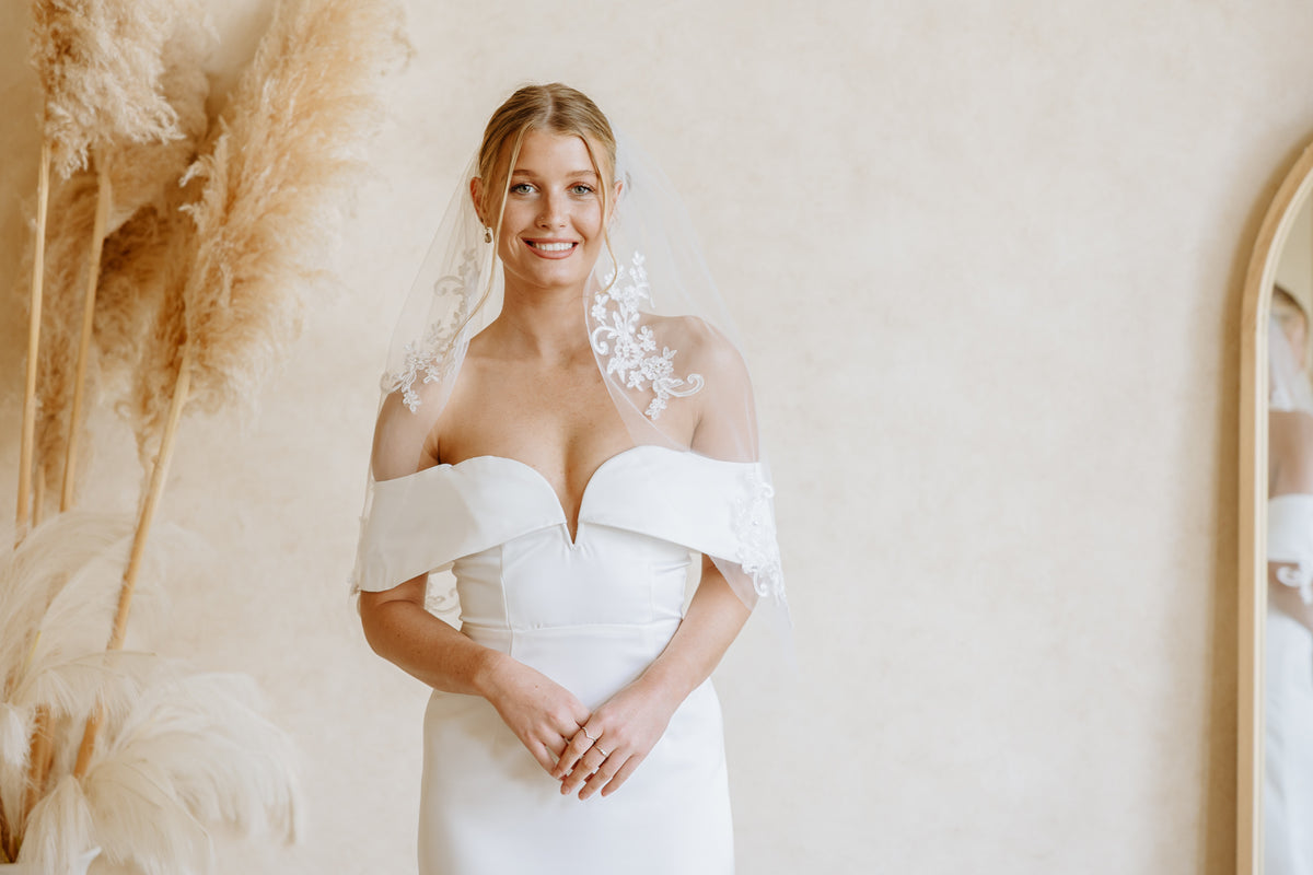 A woman wearing a white off-the-shoulder wedding dress with lace applique at the neckline and a sheer lace veil, standing indoors against a warm beige wall with dried pampas grass decor.