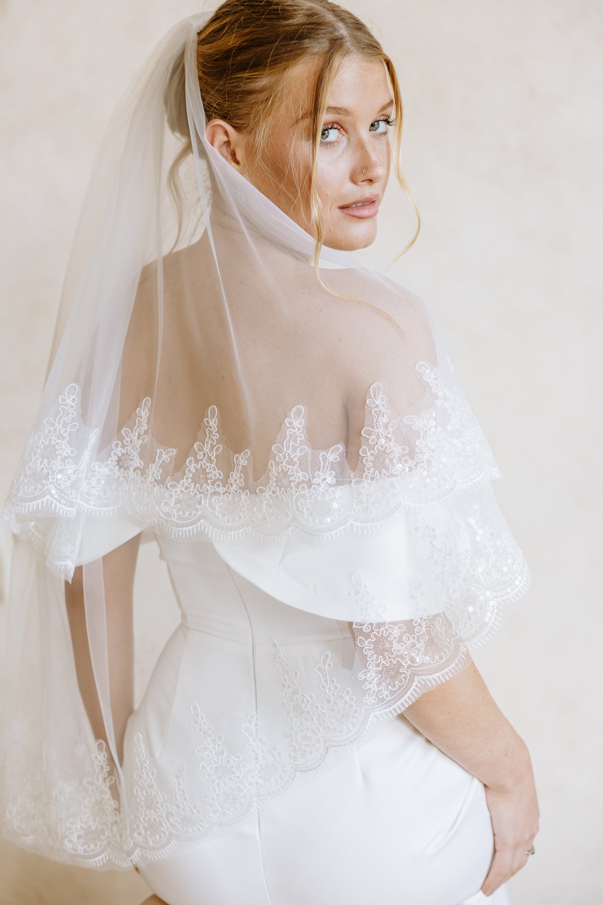 A bride wearing a white wedding dress with a sheer off-the-shoulder lace veil draped over her shoulders, photographed against a soft beige studio background.