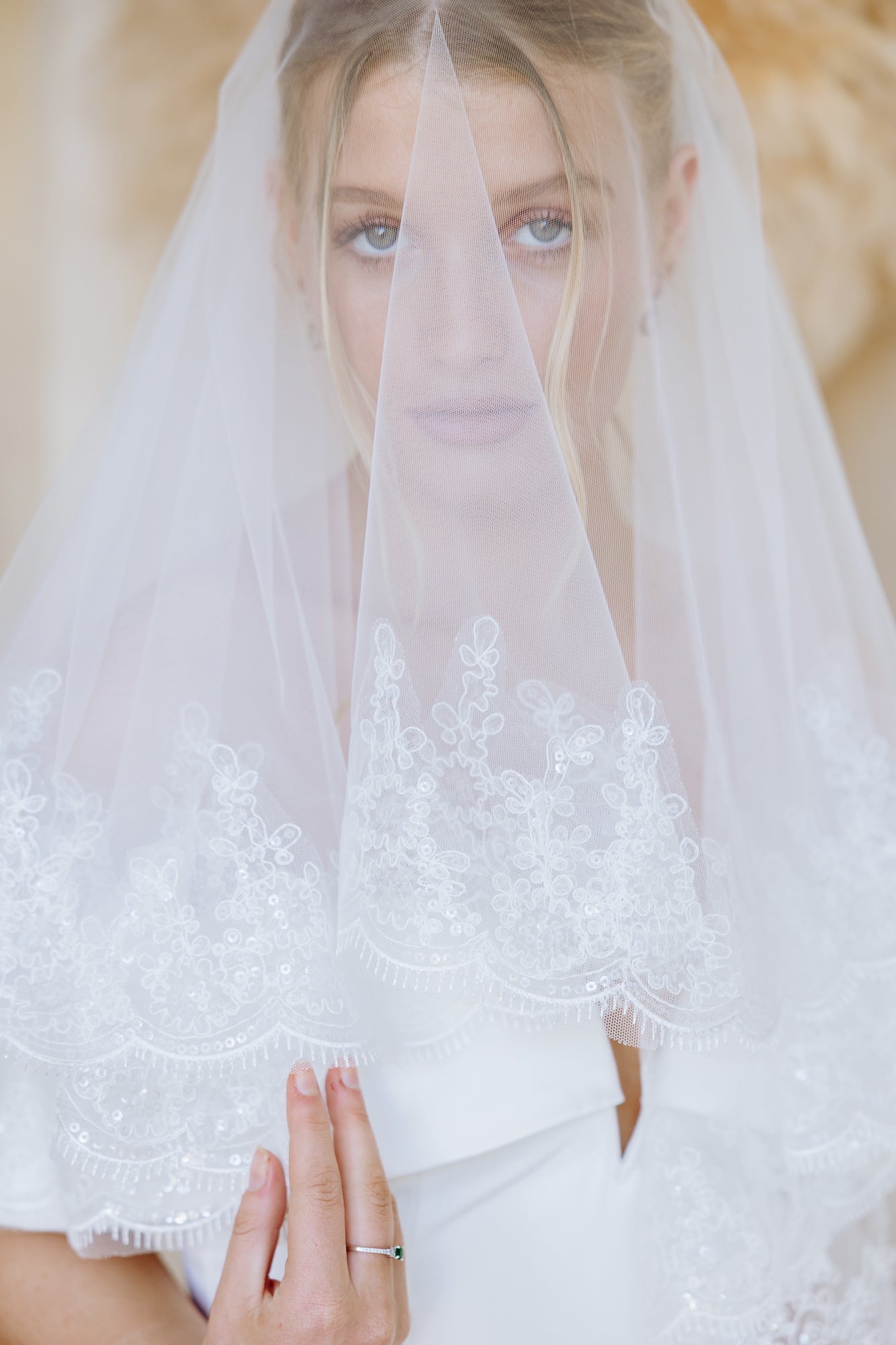 A bride wearing a white wedding dress with a sheer off-the-shoulder lace veil draped over her shoulders, photographed against a soft beige studio background.