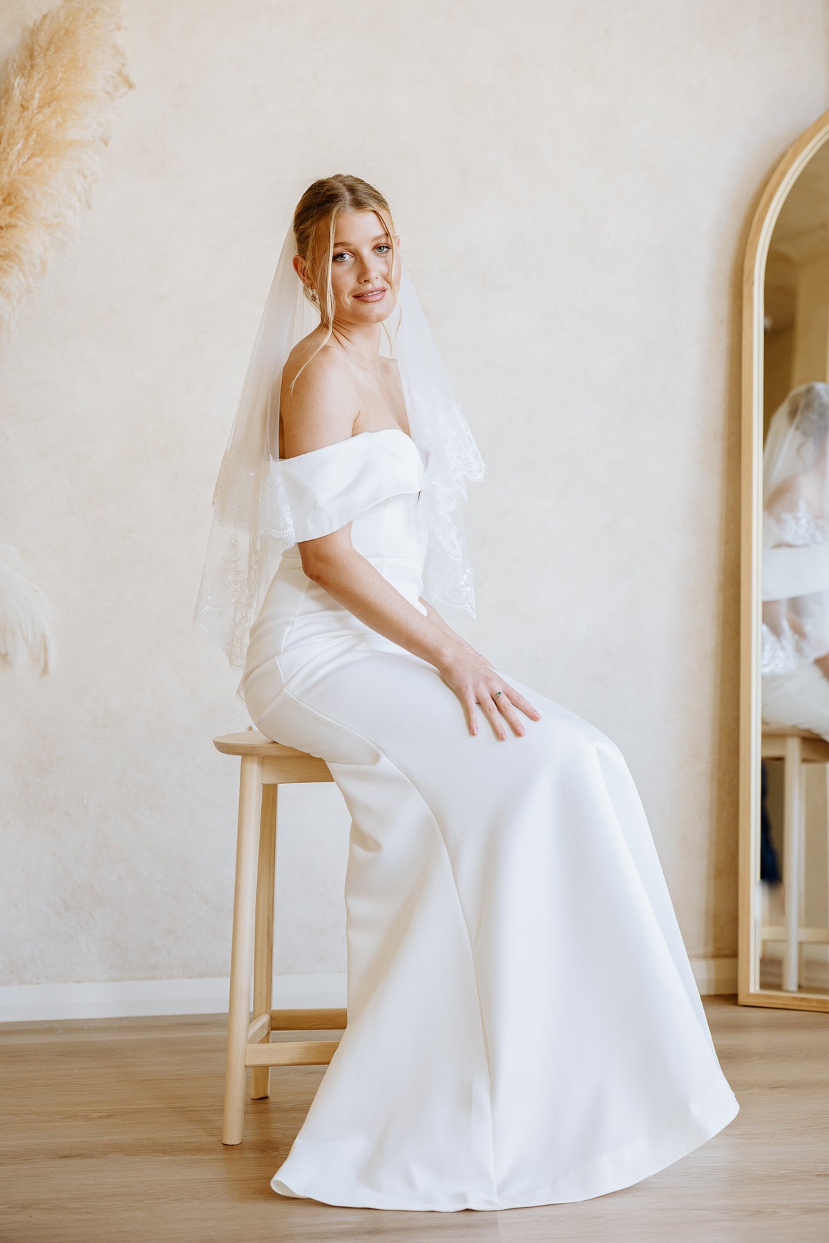 A woman wearing a white off-the-shoulder wedding dress and veil sits on a wooden stool in a softly lit indoor room with a mirror and neutral wall background.