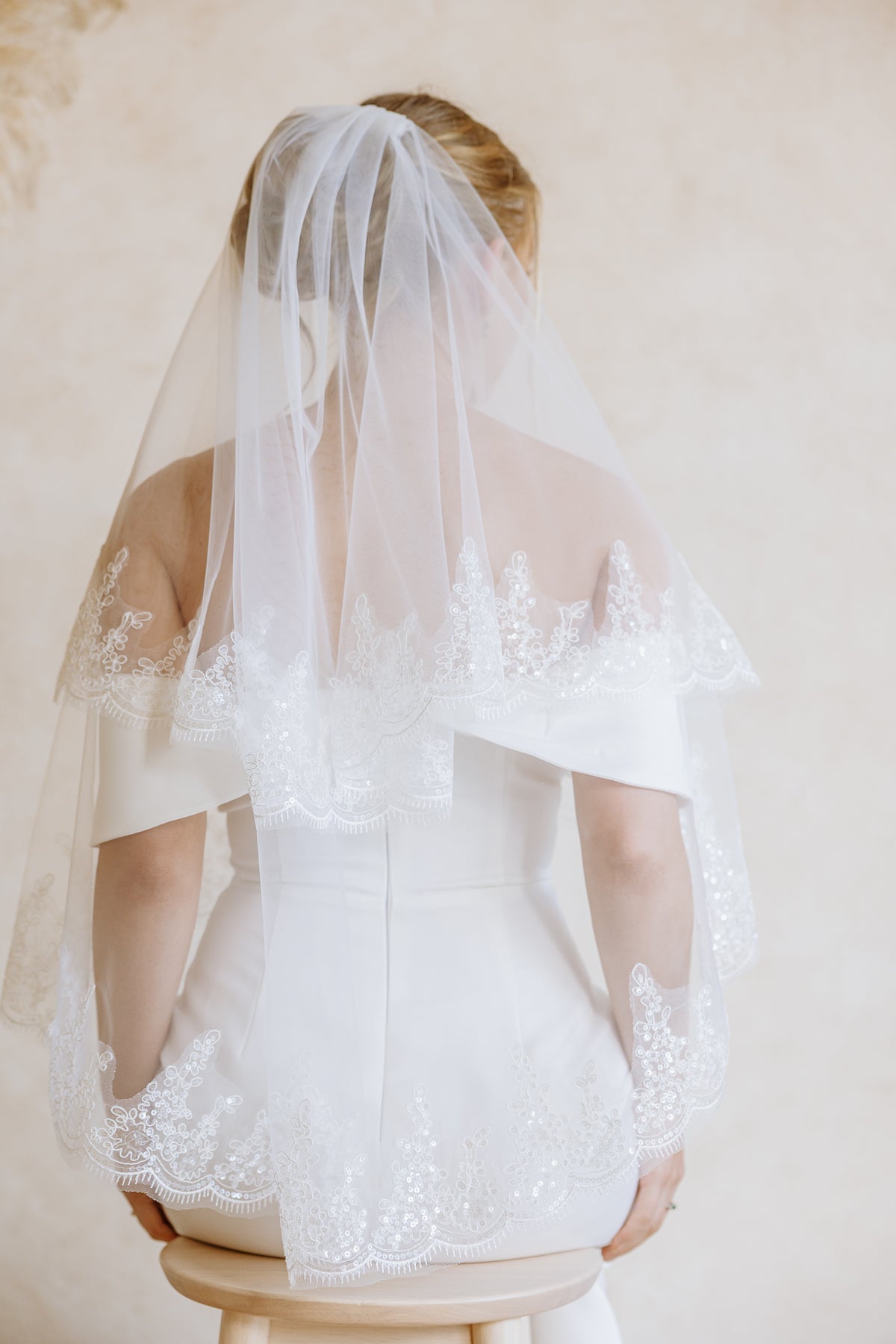A bride viewed from behind wearing a white wedding dress and a long sheer tulle veil with floral lace trim, seated on a small stool against a soft beige studio background.