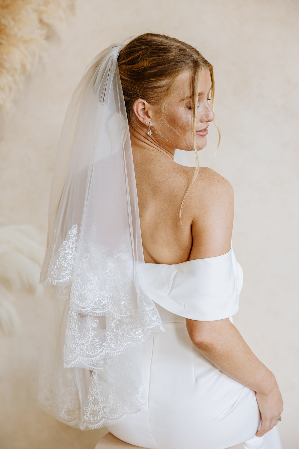 A bride wearing a white off-the-shoulder wedding dress and a long sheer tulle veil with lace trim, photographed from the back in a softly lit indoor setting.