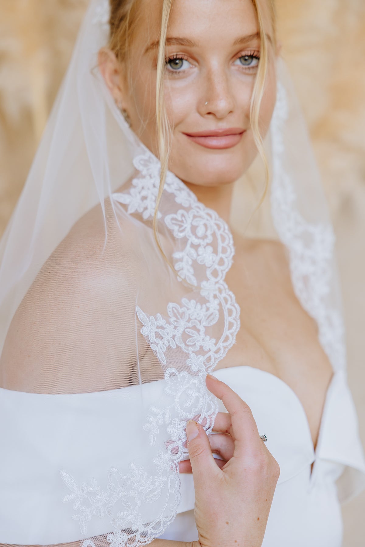 A close-up of a bride wearing a white off-the-shoulder wedding dress and a sheer white veil with ornate floral lace trim, holding the lace against her shoulder.