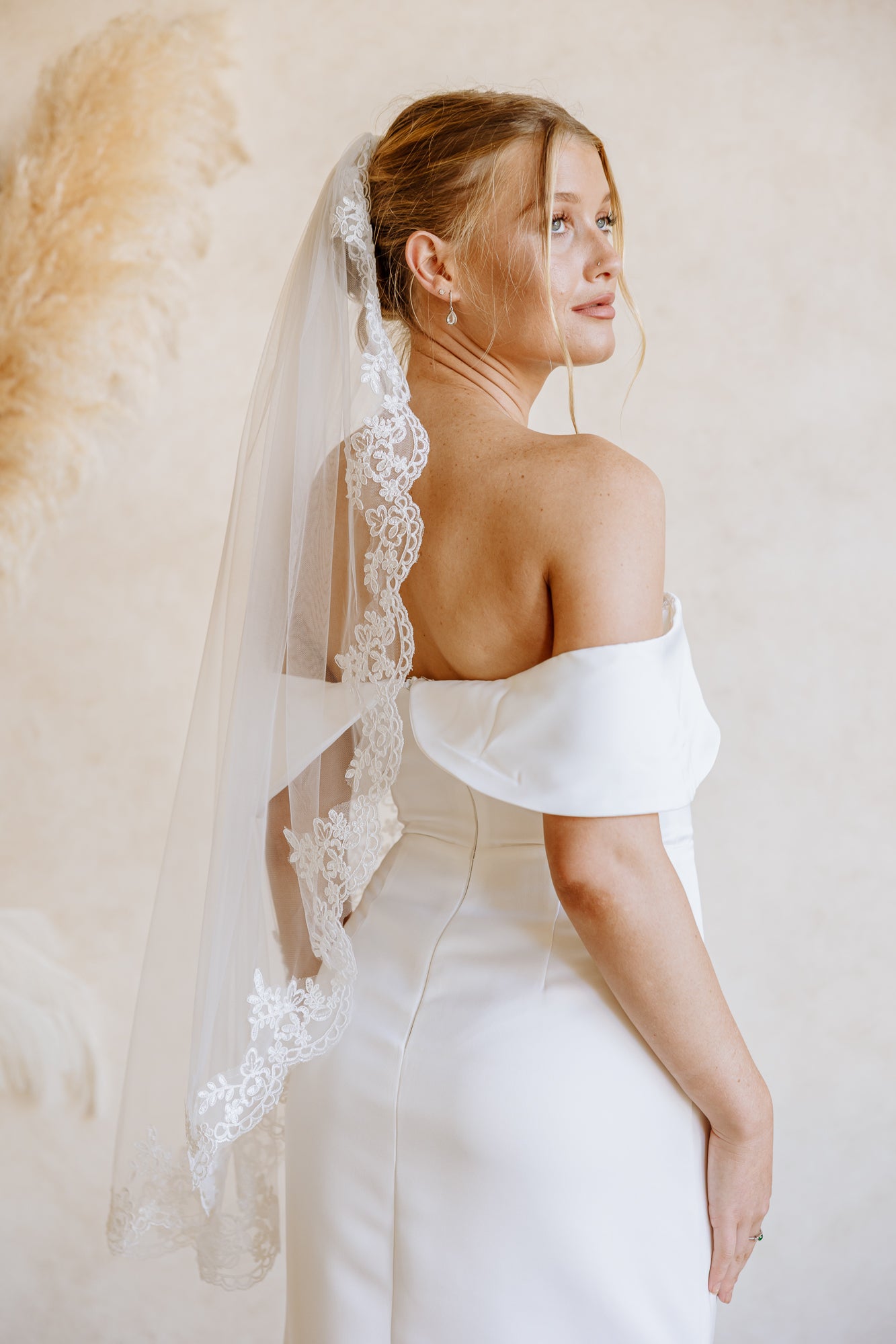 A close-up of a bride wearing a white off-the-shoulder wedding dress and a sheer white veil with ornate floral lace trim, holding the lace against her shoulder.
