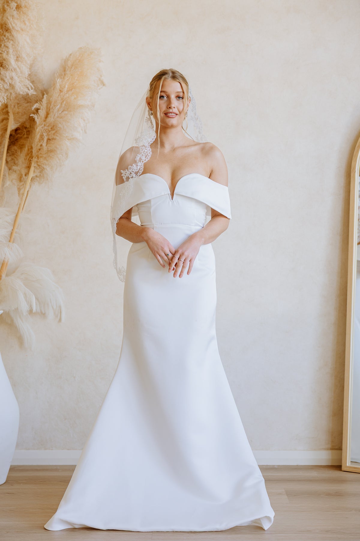 A woman wearing a white off-the-shoulder mermaid-style wedding gown with a matching veil, standing in a softly lit indoor room with beige walls and dried pampas grass decor.