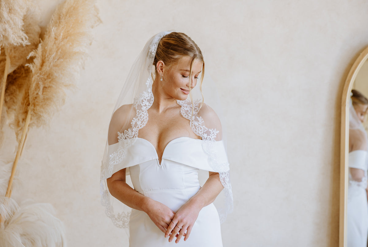 A bride wearing an off-the-shoulder white wedding dress with lace trim and a matching veil, standing indoors against a warm beige wall with dried pampas grass and a mirror visible.