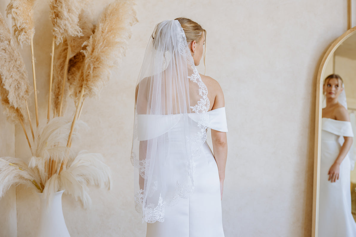 A woman wearing a white off-the-shoulder wedding dress and long sheer lace-trim veil, standing with her back to the camera in a softly lit room. A mirror on the right reflects her front, and dried pampas grass decor appears on the left.
