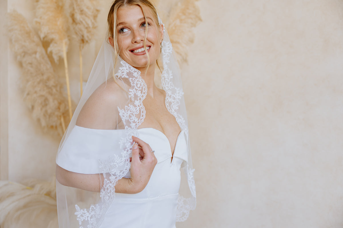 A smiling blonde woman wearing a white off-the-shoulder wedding dress and a sheer white lace-trimmed veil, posed against a soft beige wall with dried pampas grass decor.