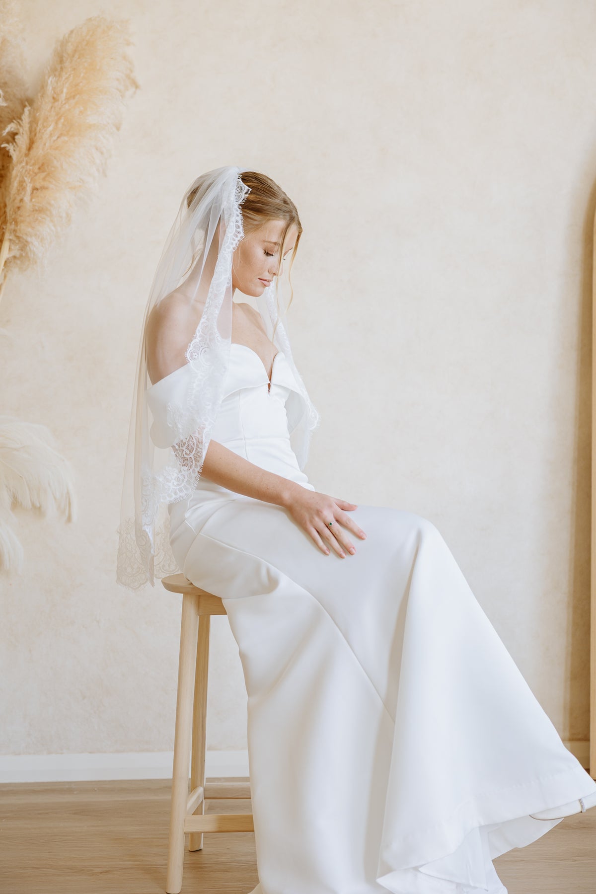 A woman in a white off-the-shoulder wedding dress with a sheer lace-trimmed veil sits on a wooden stool in a softly lit indoor setting with a textured beige wall and dried pampas grass decor.