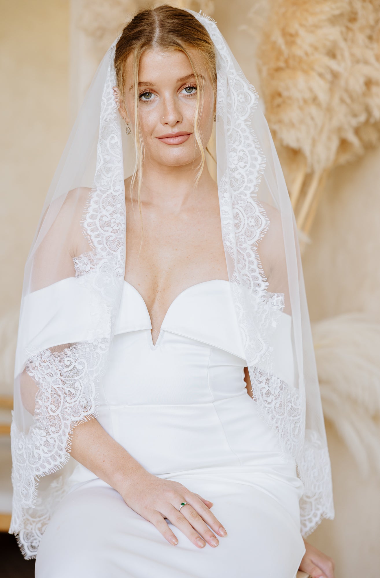 A woman in a white off-the-shoulder wedding dress with a sheer lace-trimmed veil sits on a wooden stool in a softly lit indoor setting with a textured beige wall and dried pampas grass decor.