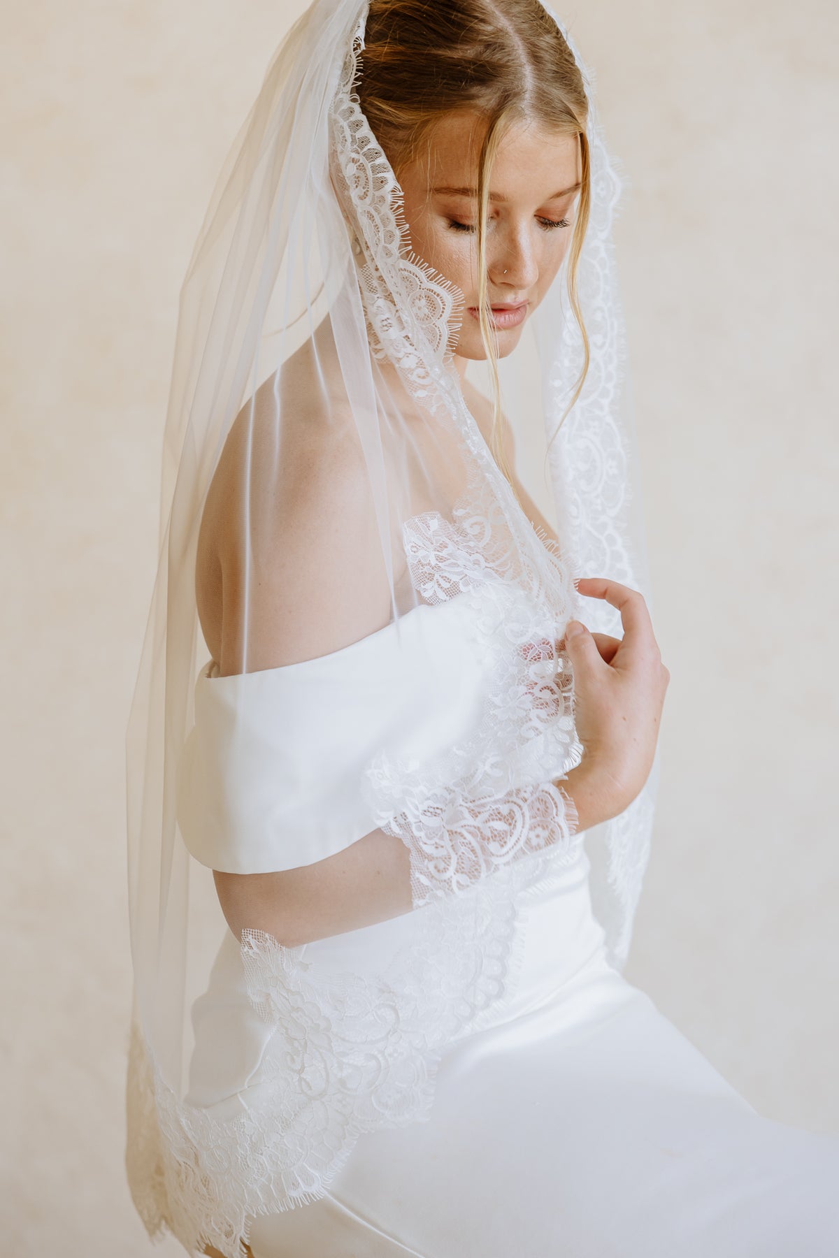 A bride wearing a white off-the-shoulder wedding dress with lace applique and a matching sheer veil, posed against a soft beige background.