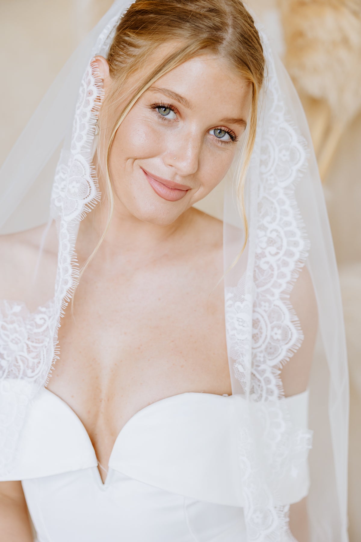 A close-up portrait of a bride wearing a white strapless wedding dress and a sheer white lace-trimmed veil, smiling softly against a warm neutral background.