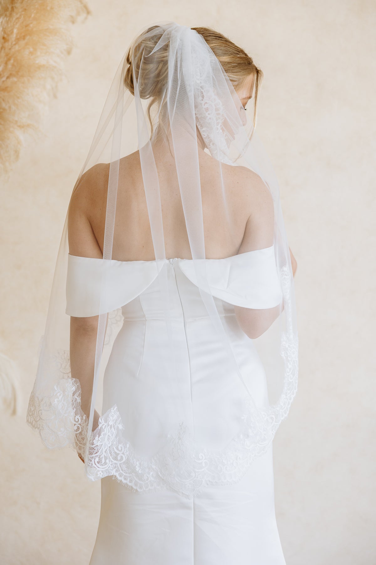 A bride viewed from behind wearing a white off-the-shoulder wedding dress and a long sheer white tulle veil with lace trim, posed against a soft beige textured backdrop.