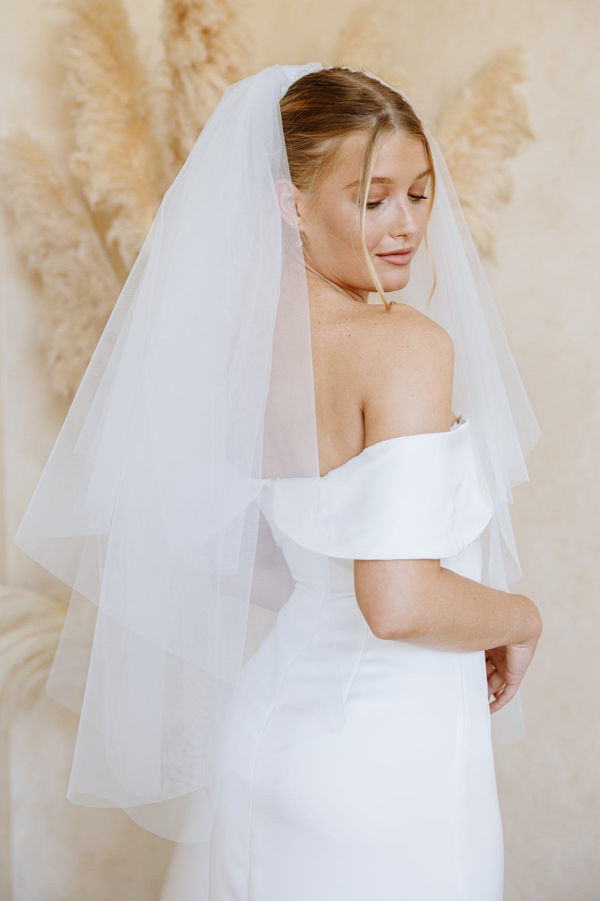 A woman wearing a white off-the-shoulder wedding dress and a long sheer tulle bridal veil, posed against a warm beige background with dried pampas grass.