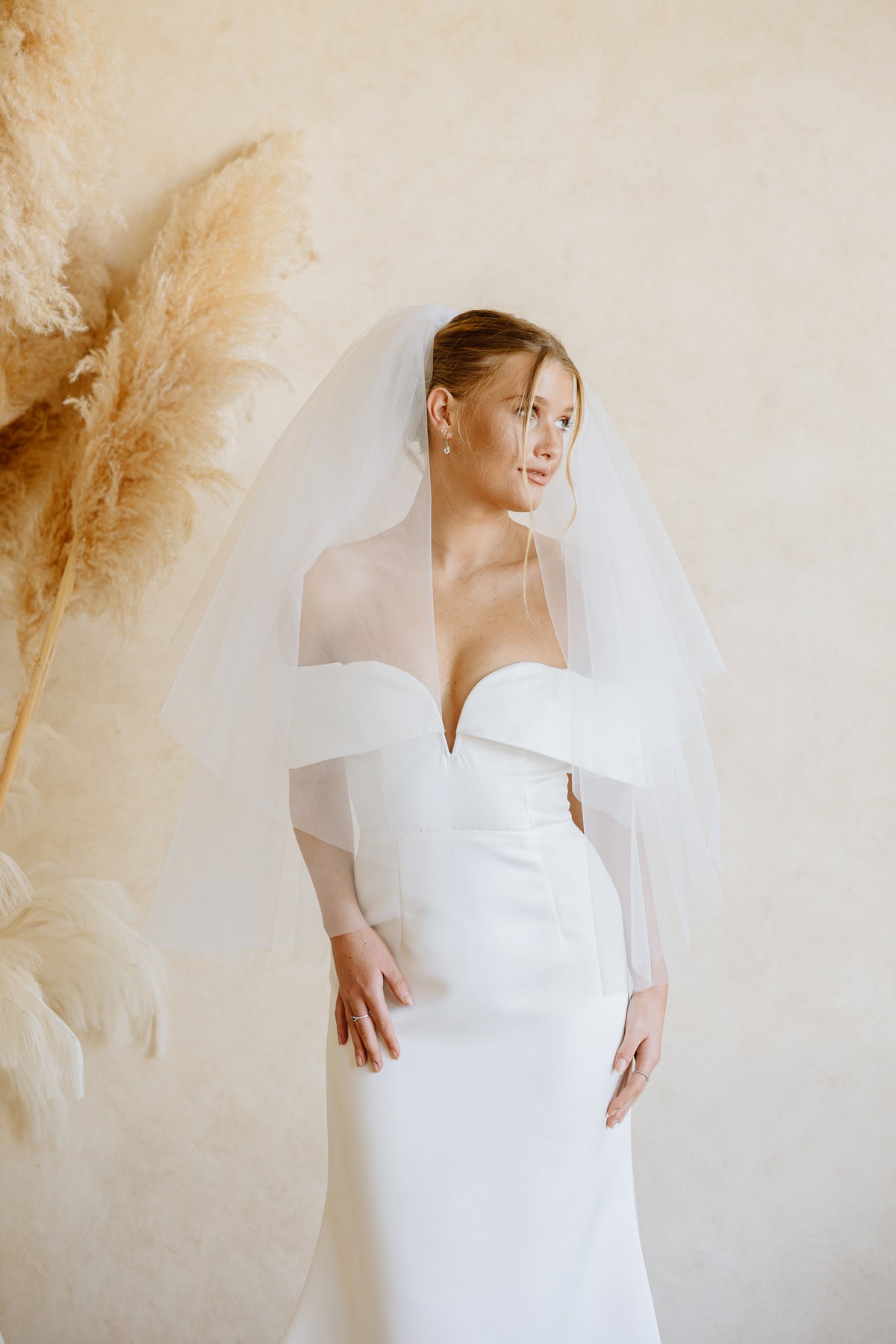 A woman wearing a white off-the-shoulder wedding dress with a fingertip length sheer veil, standing against a warm beige wall with pampas grass decor on the left.