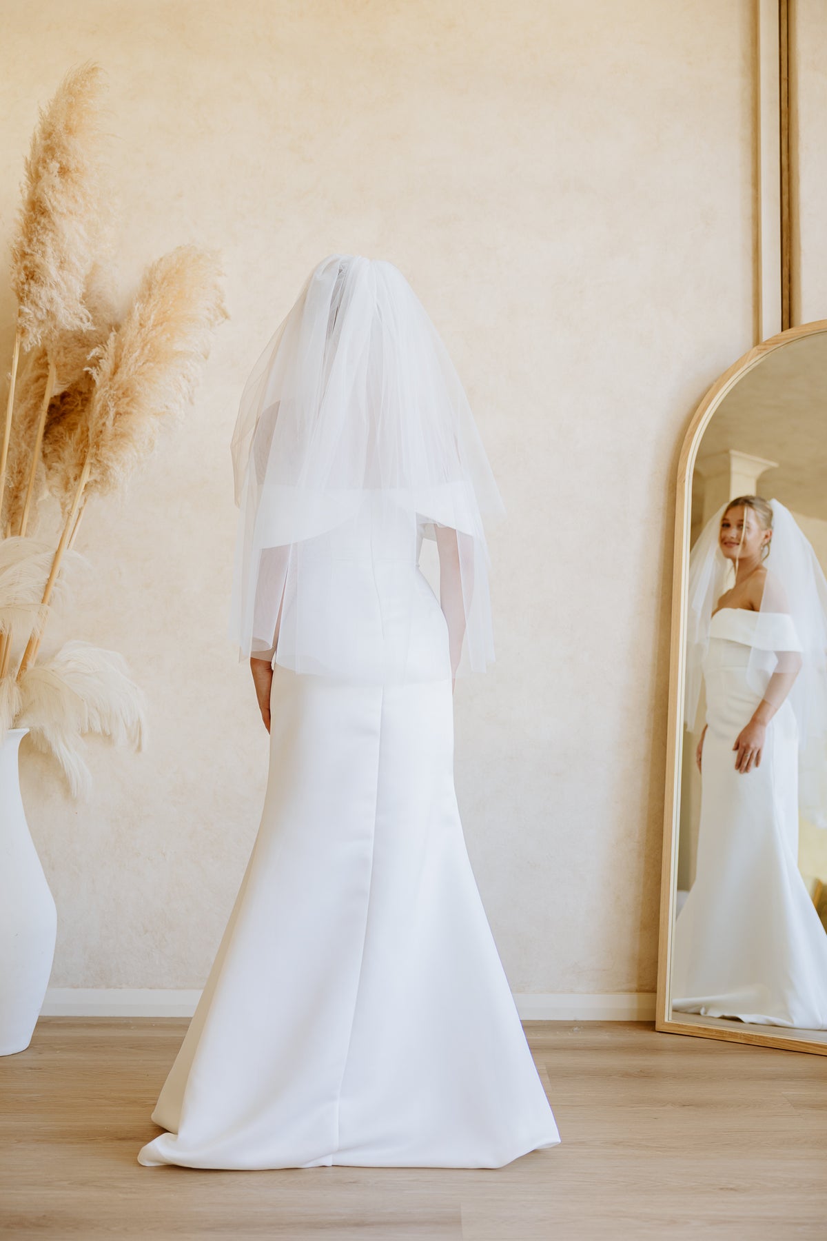 A bride wears a white wedding dress and a tulle veil, with a mirror on the right reflecting her side and dried pampas grass on the left, view from the back