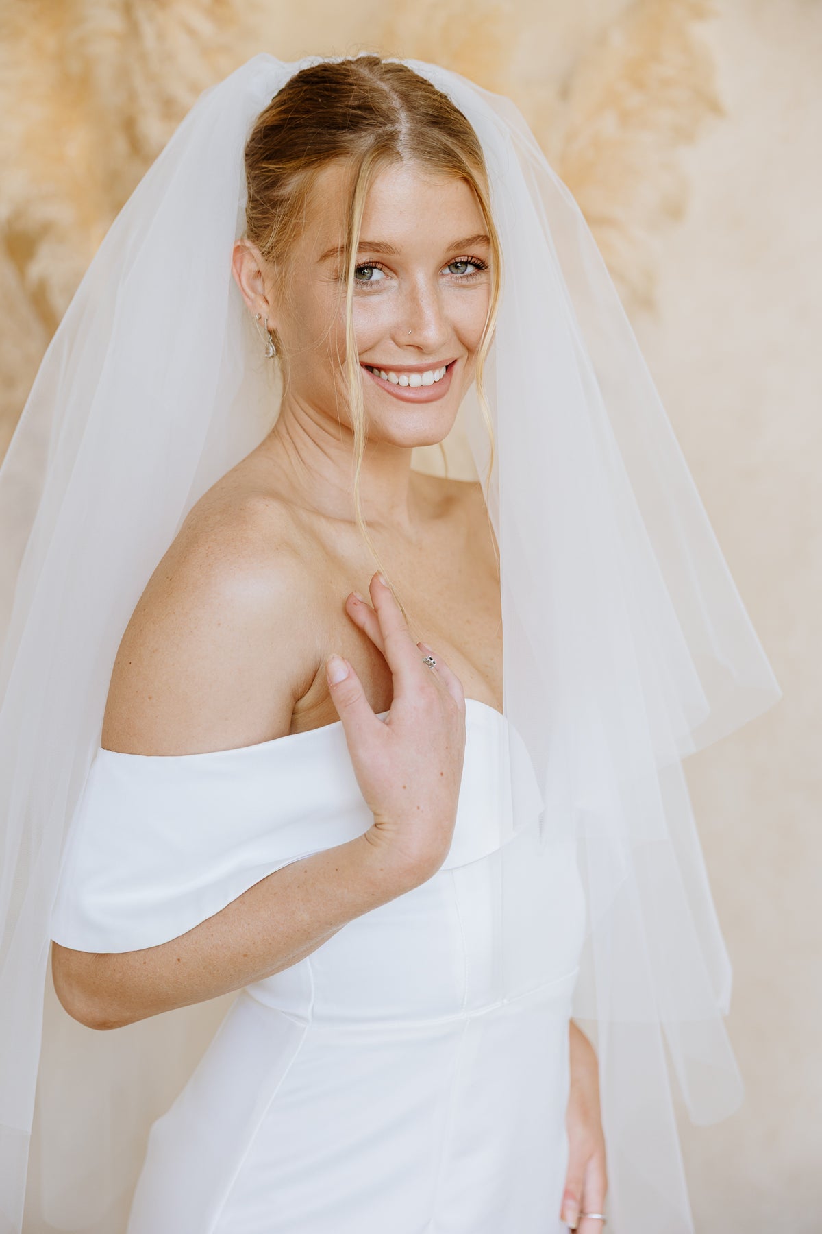 A smiling bride wearing a white off-the-shoulder wedding dress and a soft sheer veil, posed against a warm beige background with dried pampas grass.