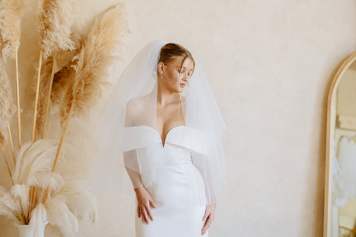 A woman wearing a white off-the-shoulder wedding dress with a sheer veil, standing indoors against a warm beige wall with pampas grass decor on the left and part of a mirror on the right.