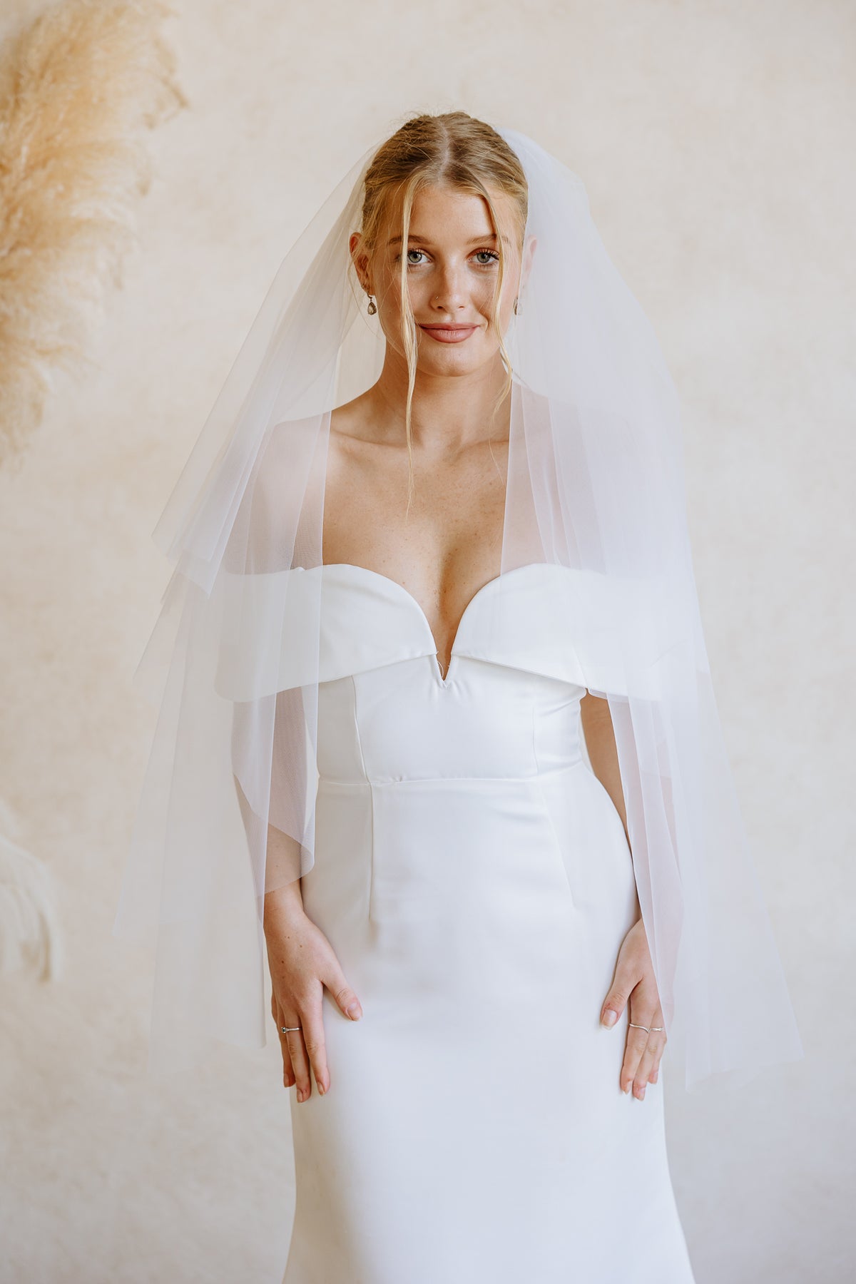 A bride wearing a white wedding dress with a sweetheart neckline and a sheer tulle veil, standing against a warm beige textured wall.