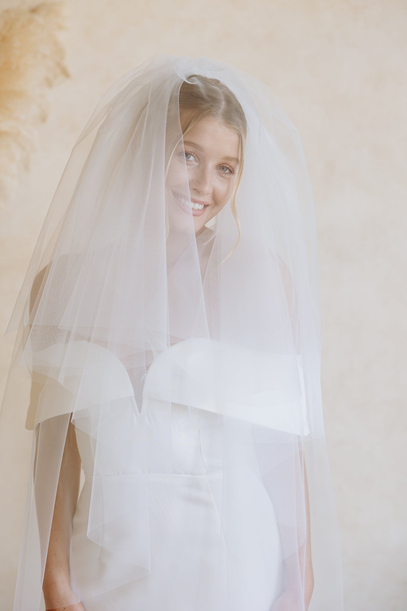 A woman wearing a white off-the-shoulder wedding dress and a long sheer tulle veil, photographed from the back against a warm beige wall with pampas grass decor.