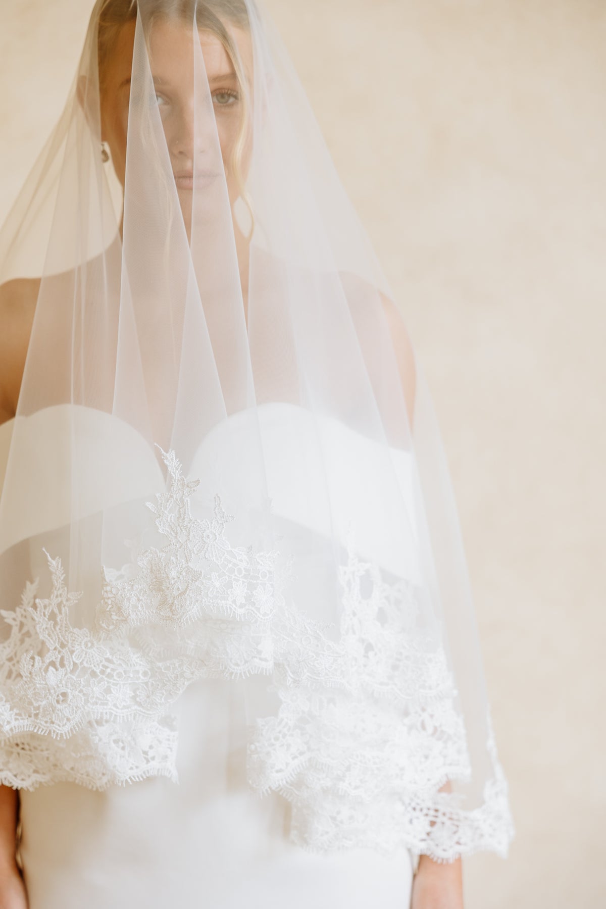 A woman wearing a white off-the-shoulder wedding dress and a long sheer veil with lace trim draped over her face