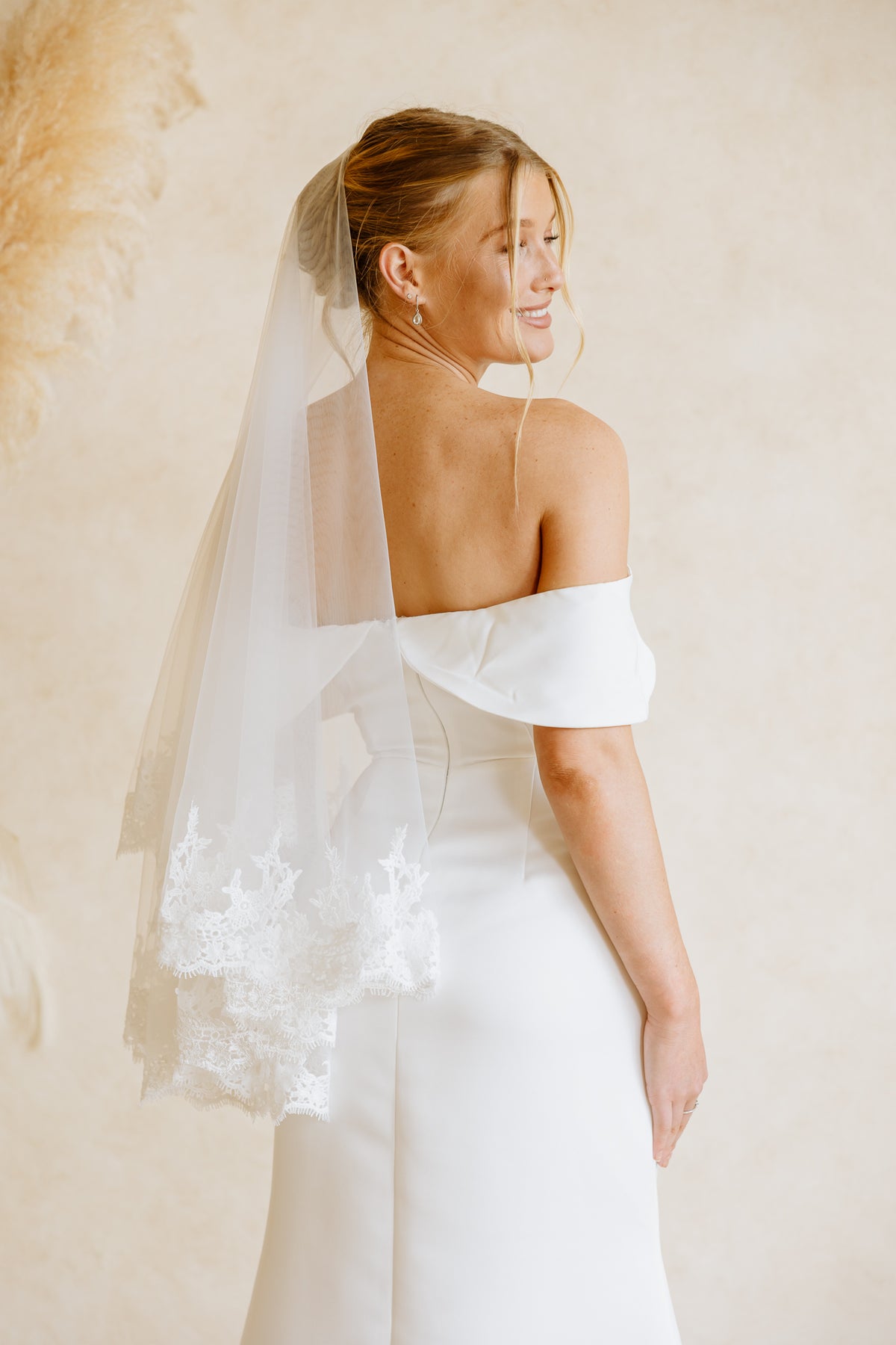 A bride smiles as she looks over her shoulder, wearing a white off-the-shoulder wedding dress and a sheer veil behind her head