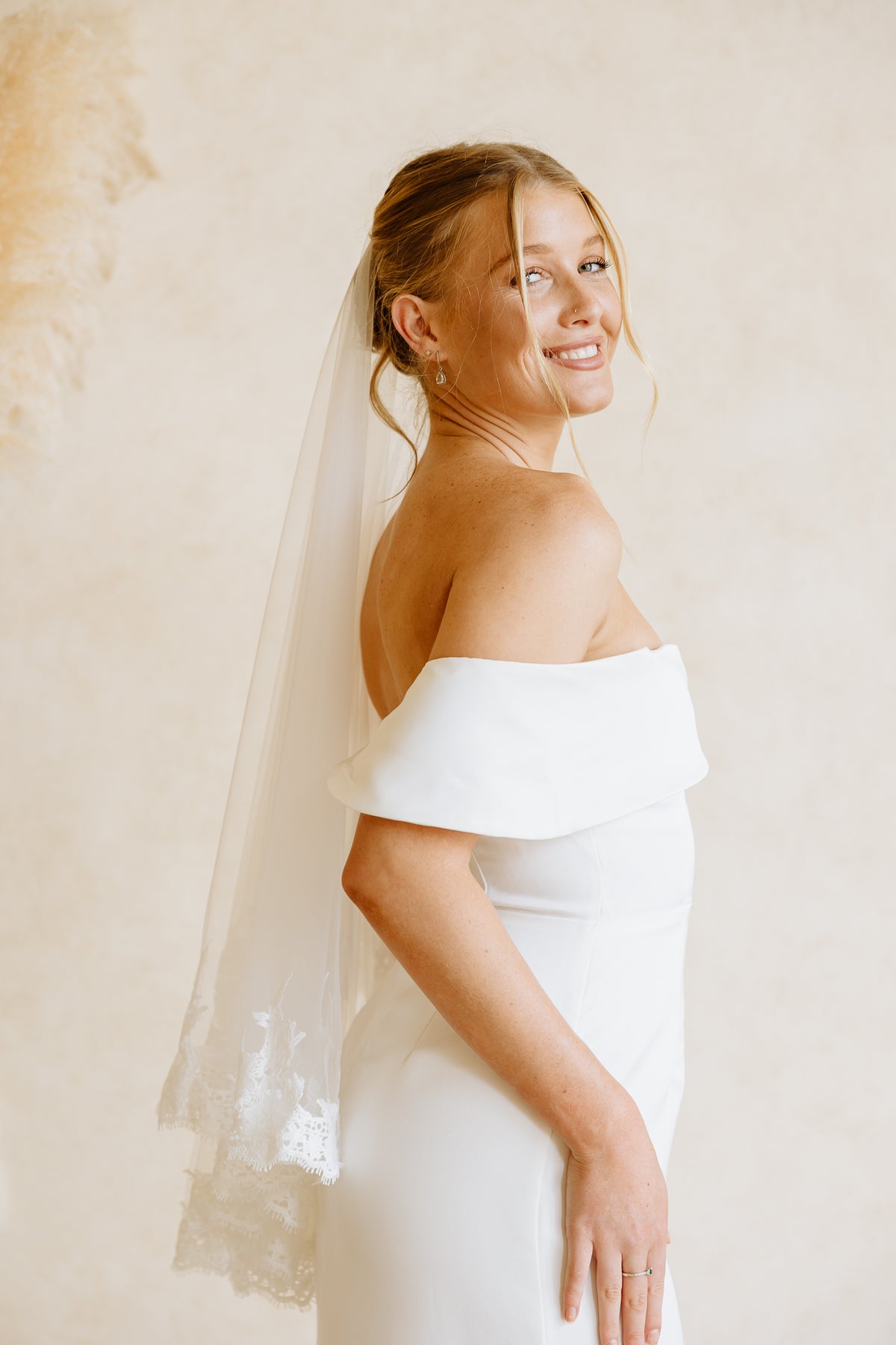 A woman wearing a white off-the-shoulder wedding dress and a lace trim veil, photographed from the back against a soft beige wall with pampas grass decor.