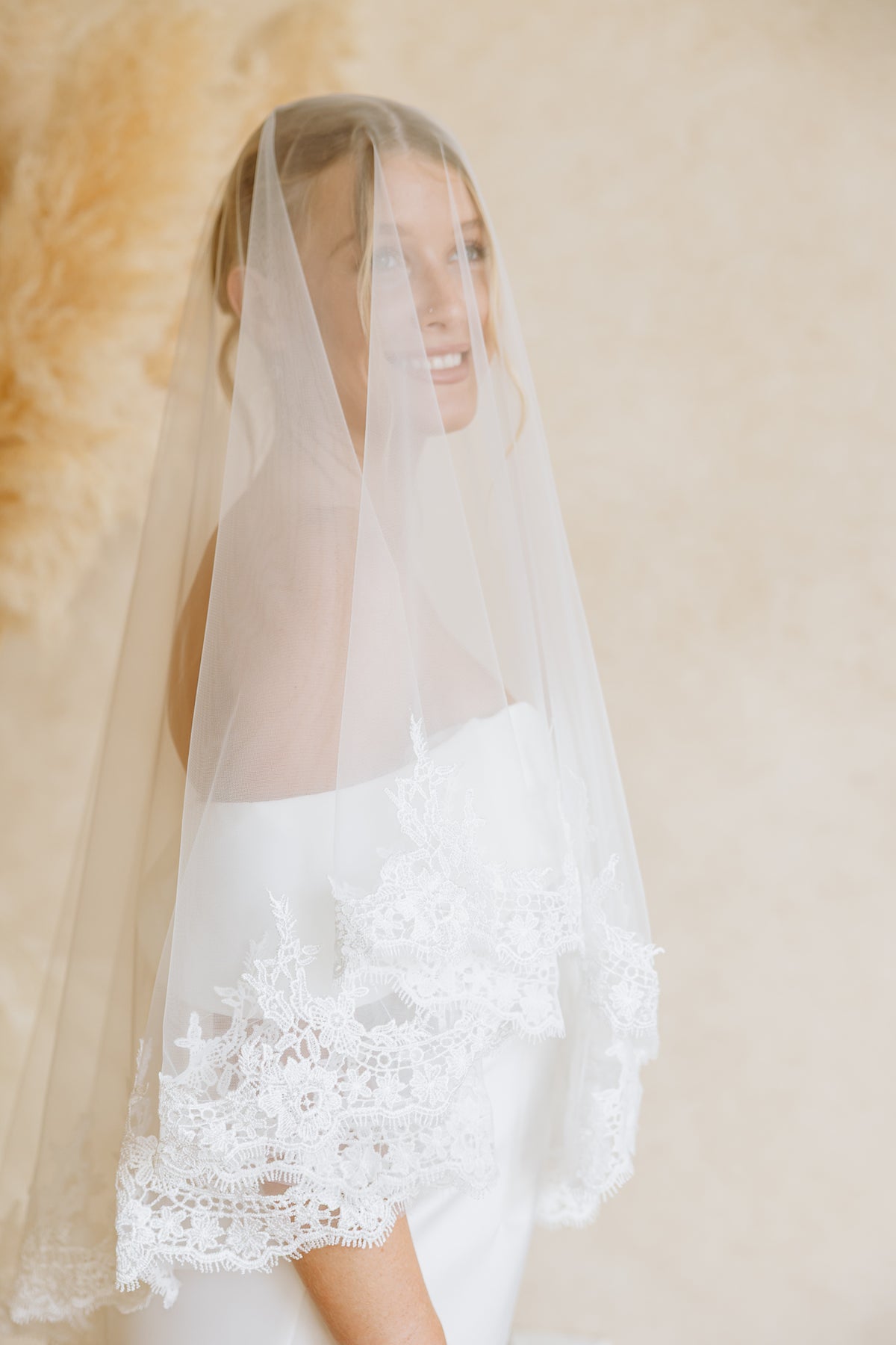 A bride smiles as she wears a sheer veil with delicate lace trim draped over her face 