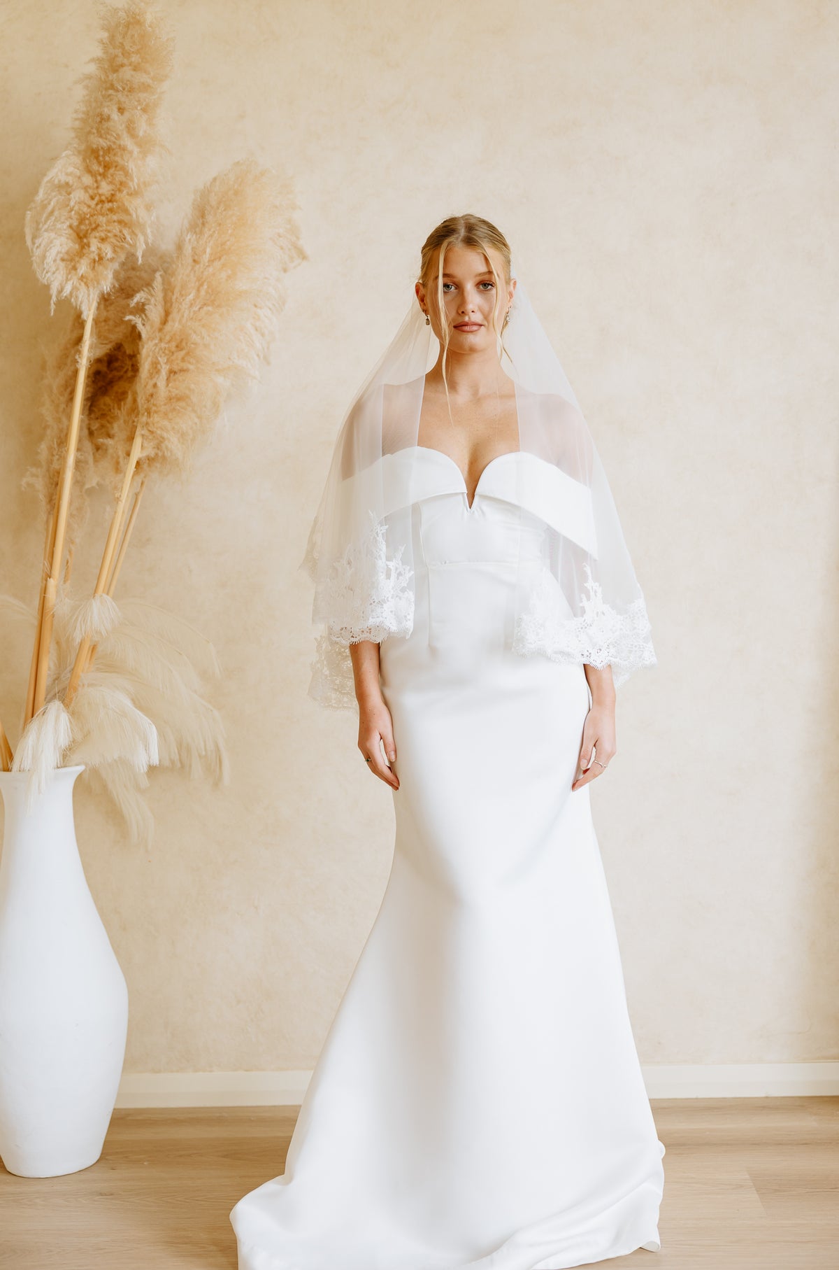 A woman wearing a white off-the-shoulder wedding dress and a long sheer veil, photographed standing in front of a soft beige wall with pampas grass decor.