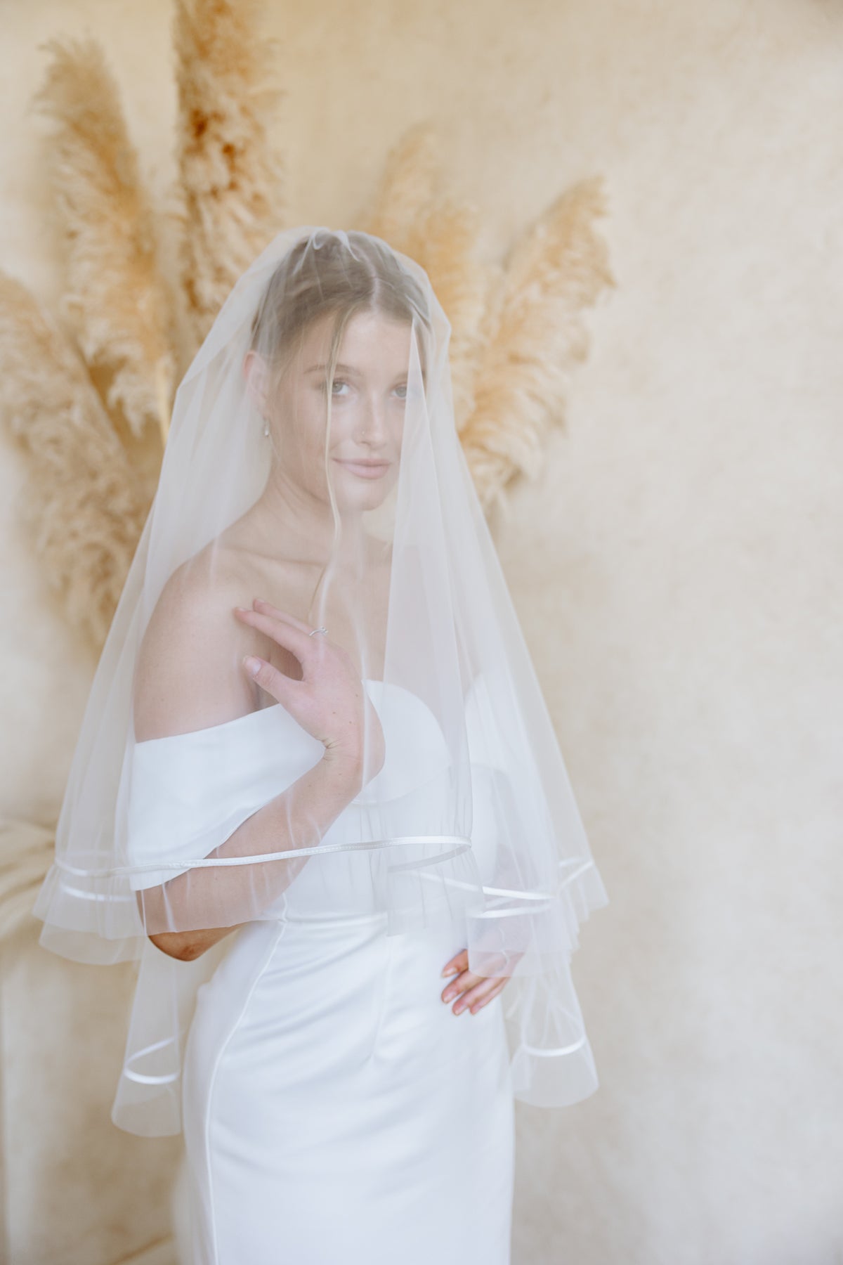 A bride in a white off-the-shoulder dress wears a sheer blusher veil with ribbon trim draped over her face, posing in front of tall, feathery pampas grass against a beige wall.