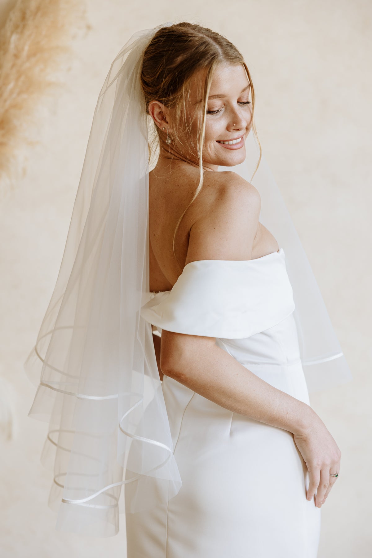 A smiling bride in an off-the-shoulder white wedding dress wears a sheer, two-tiered veil with a satin ribbon trim against a neutral beige background.