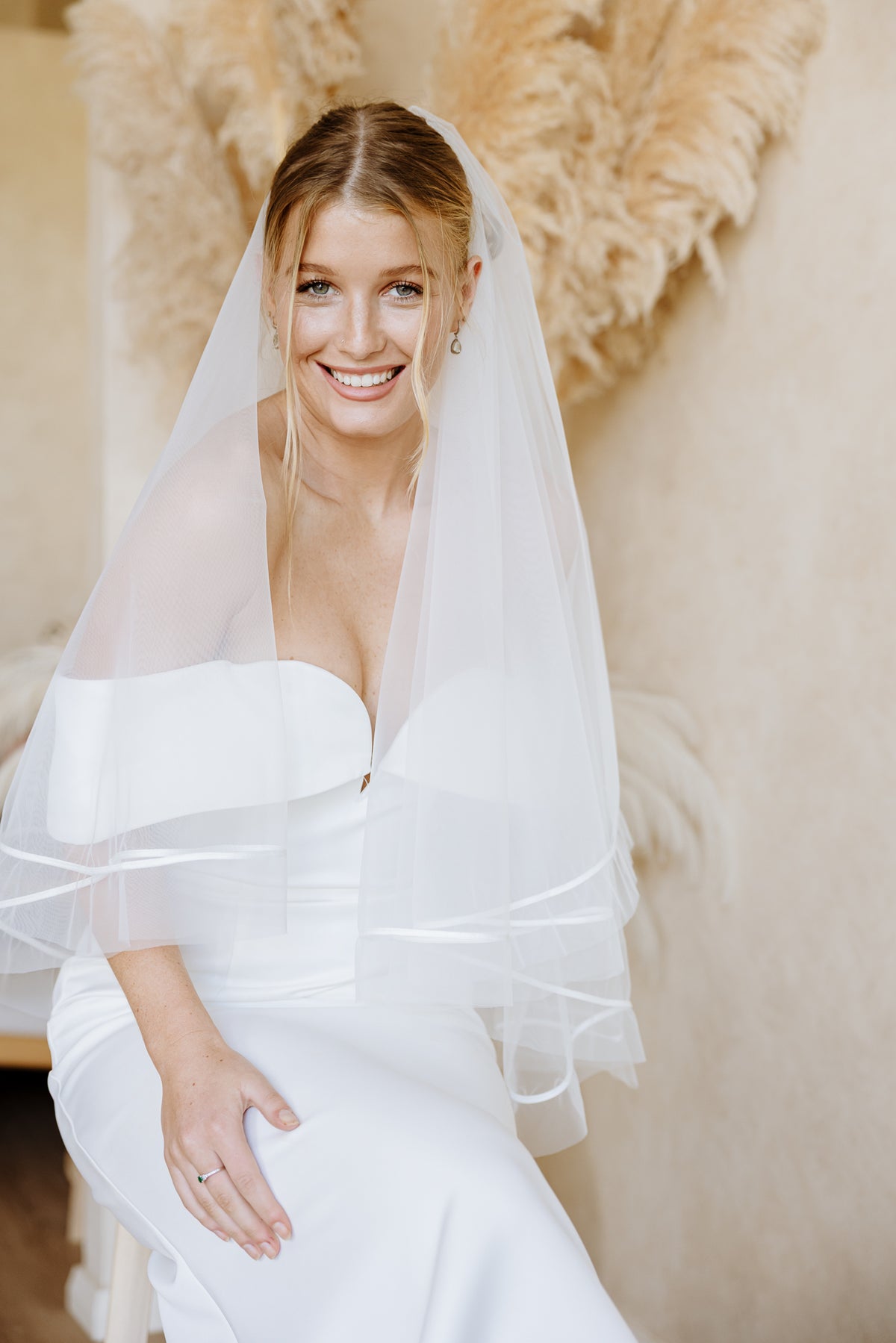 A smiling bride in an off-the-shoulder white wedding dress wears a sheer, layered veil with a satin ribbon trim against a neutral beige background.