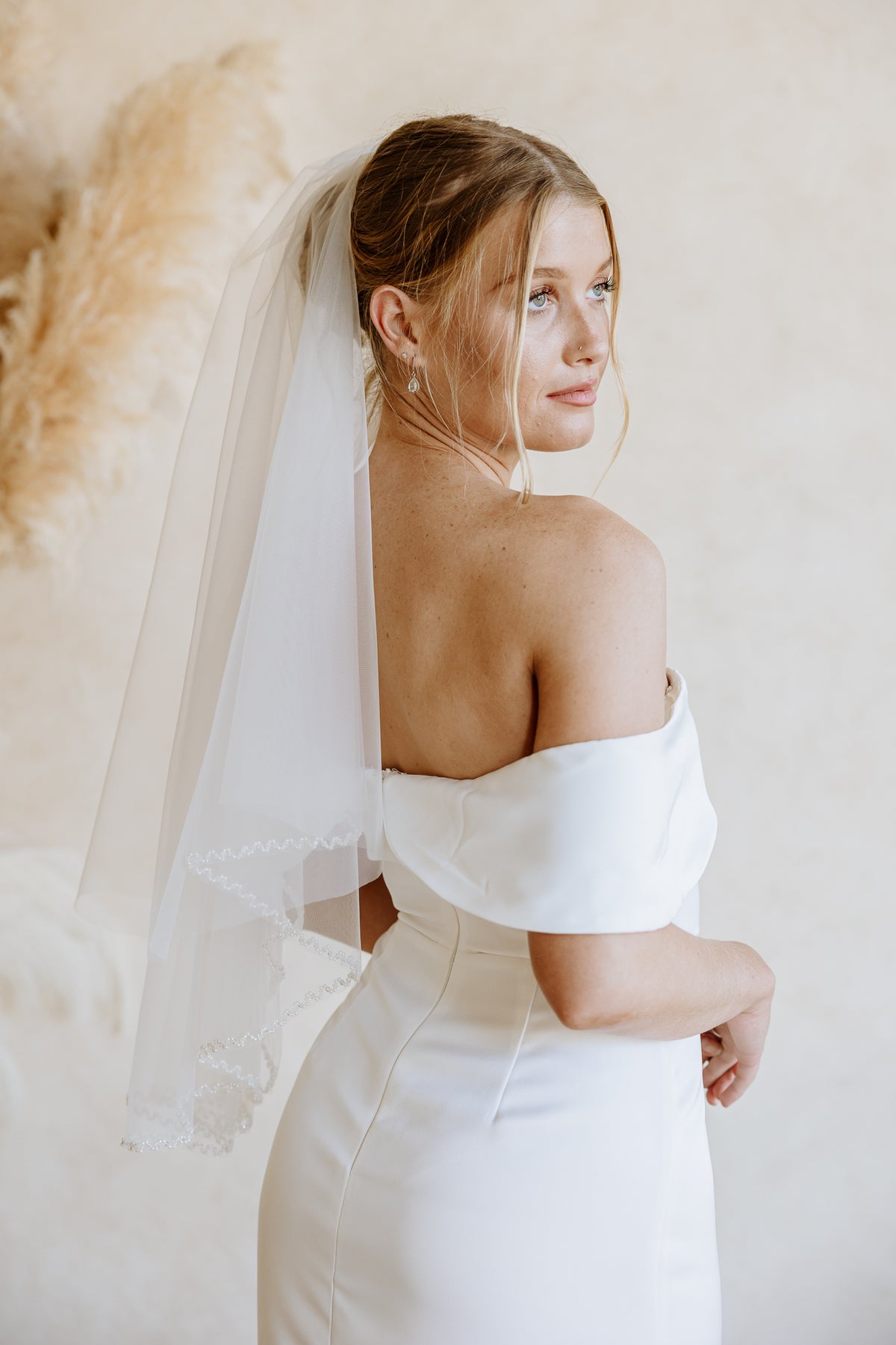 A woman wearing a white off-the-shoulder wedding dress and a long sheer veil, photographed from the back against a soft beige wall with pampas grass decor.