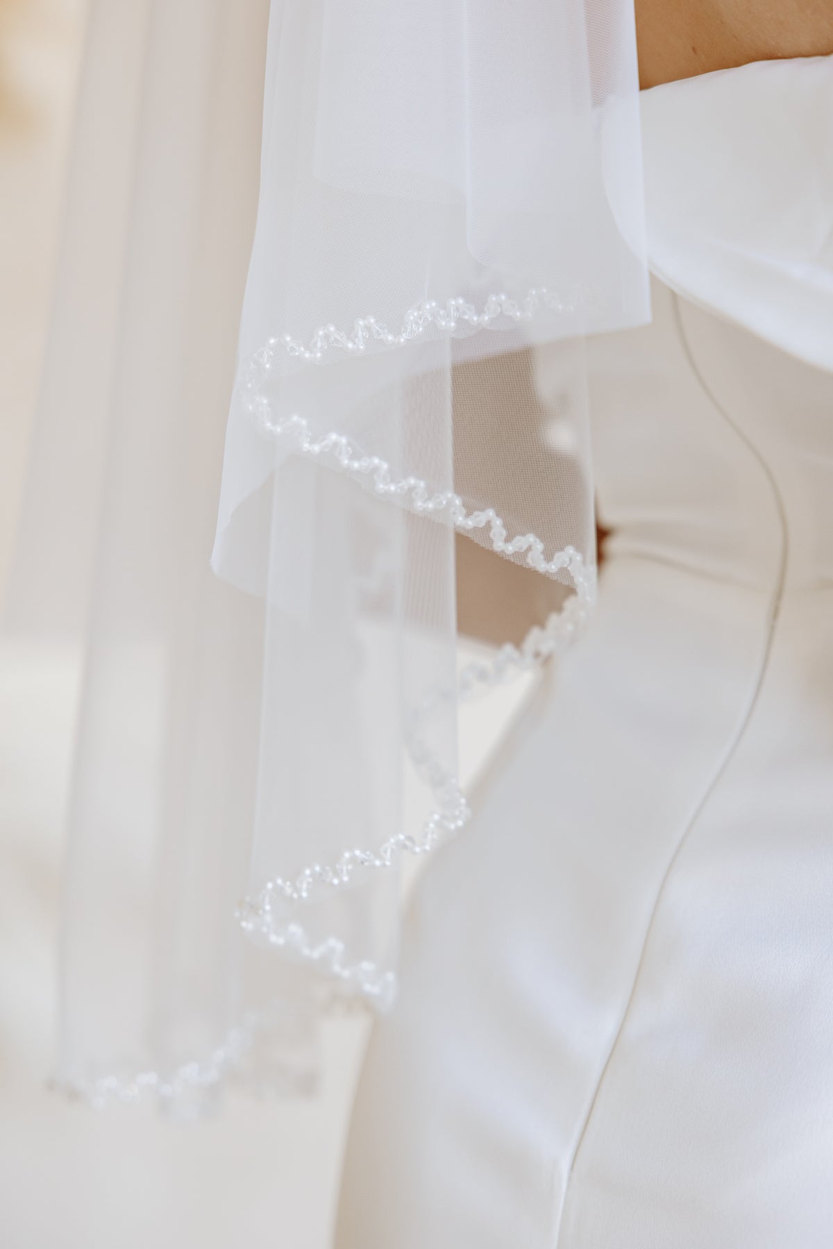 A close-up of a white bridal veil with delicate beaded scalloped edges draped over a white wedding dress.