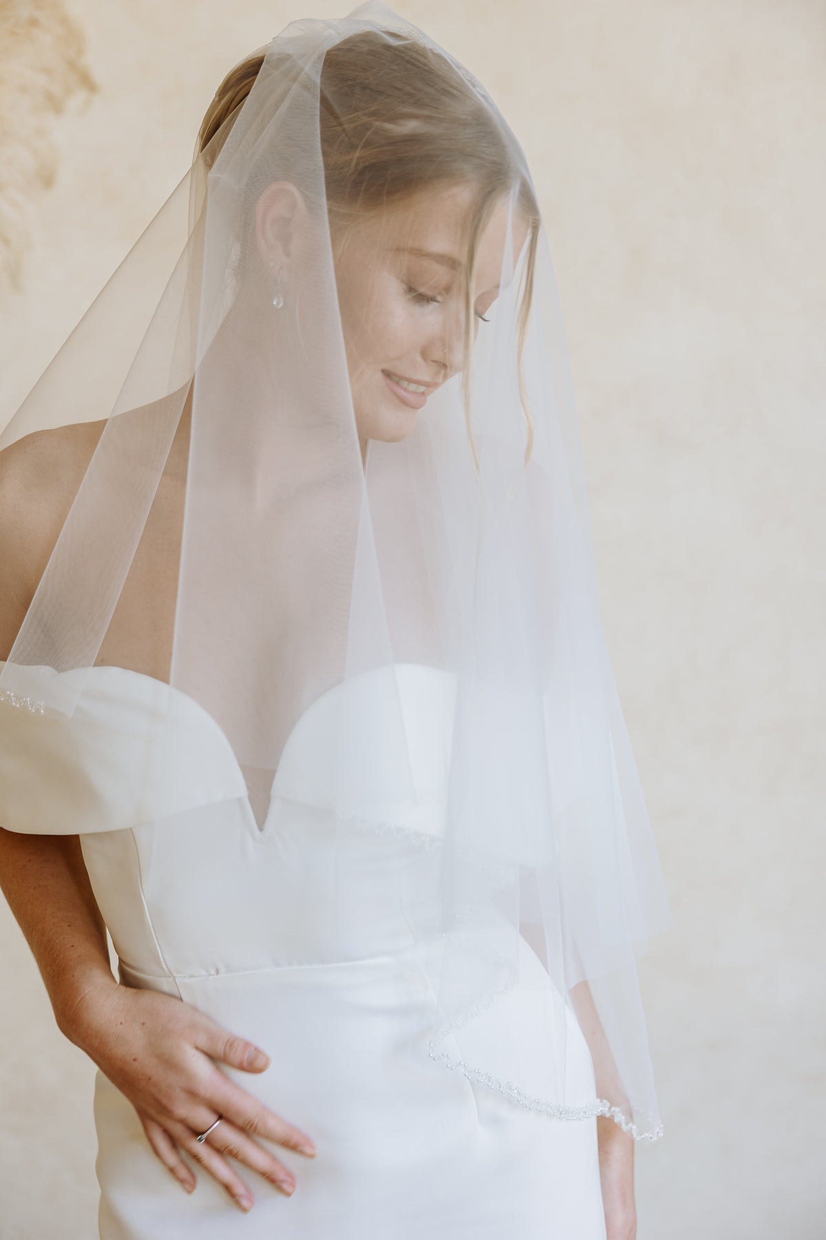 A bride wearing a sheer white tulle bridal veil and a strapless white wedding dress, shown in a soft, warm-toned portrait against a neutral beige background.