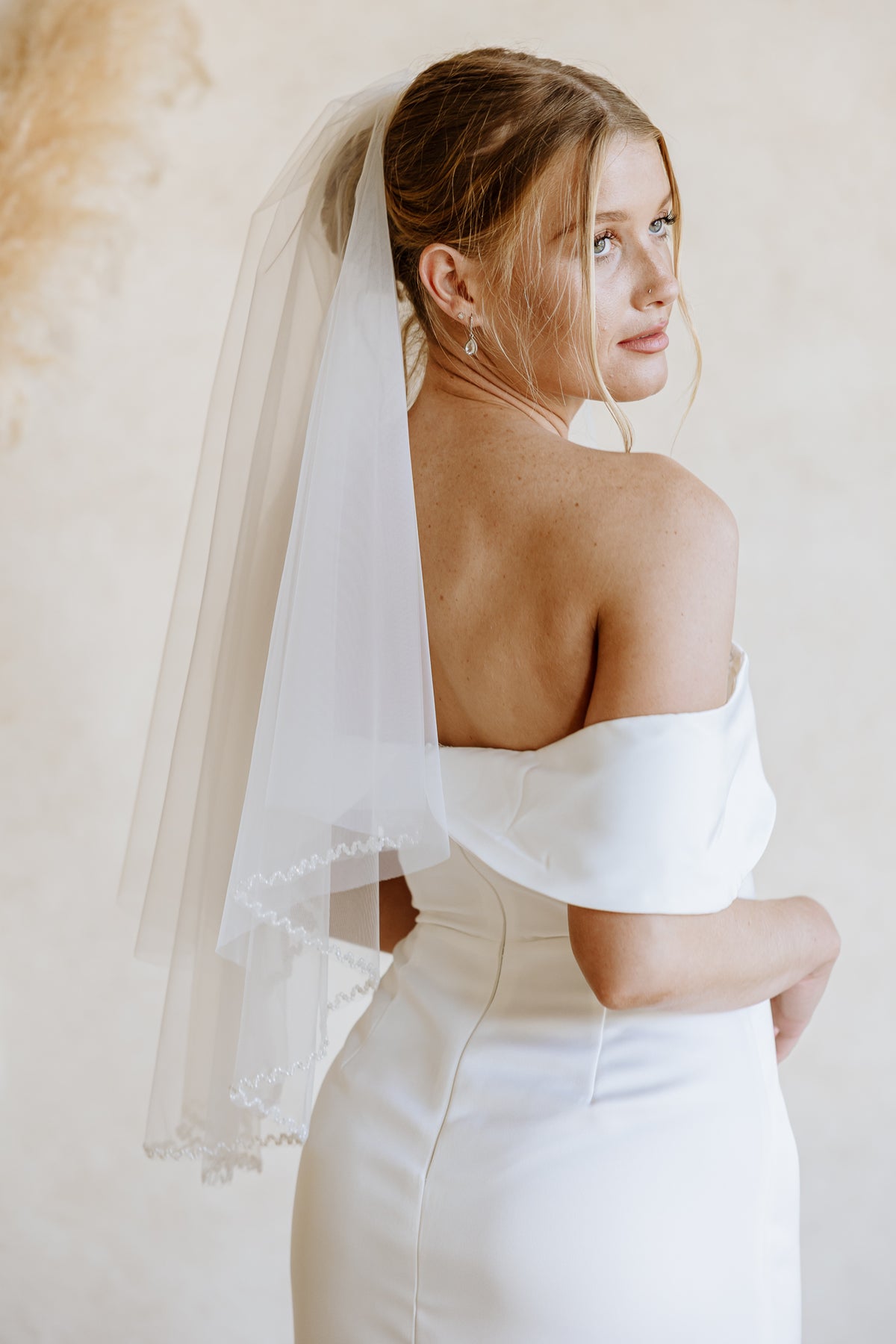 A woman wearing a white off-the-shoulder wedding dress and a long sheer tulle veil, photographed from the back against a soft beige studio background.