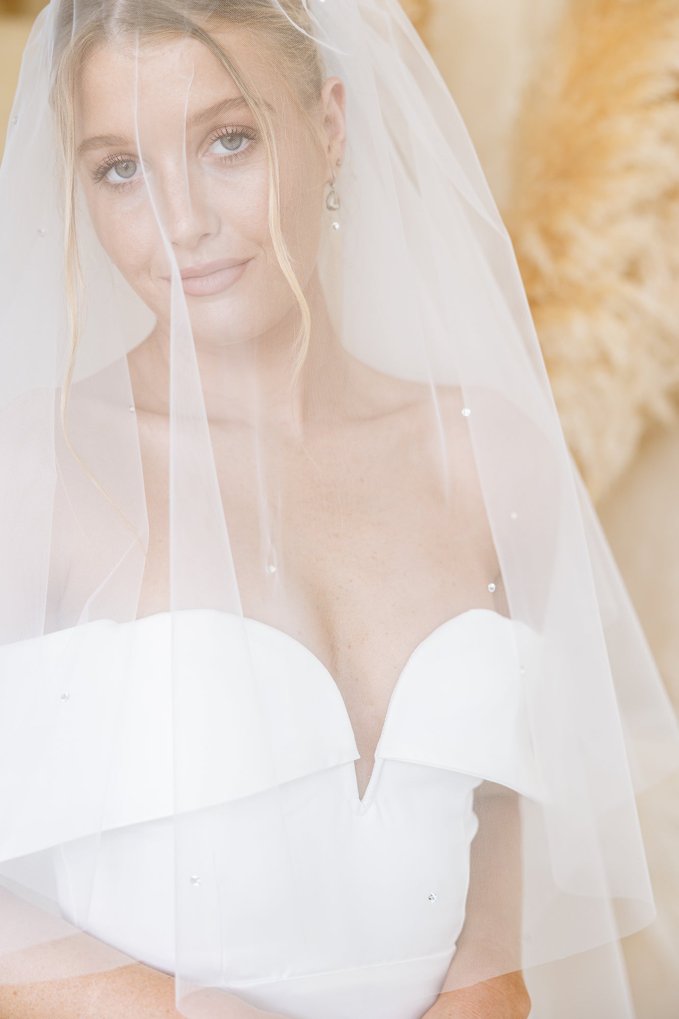 A smiling bride in a fitted white wedding dress with a sheer off-the-shoulder veil, posed against a soft beige textured wall.