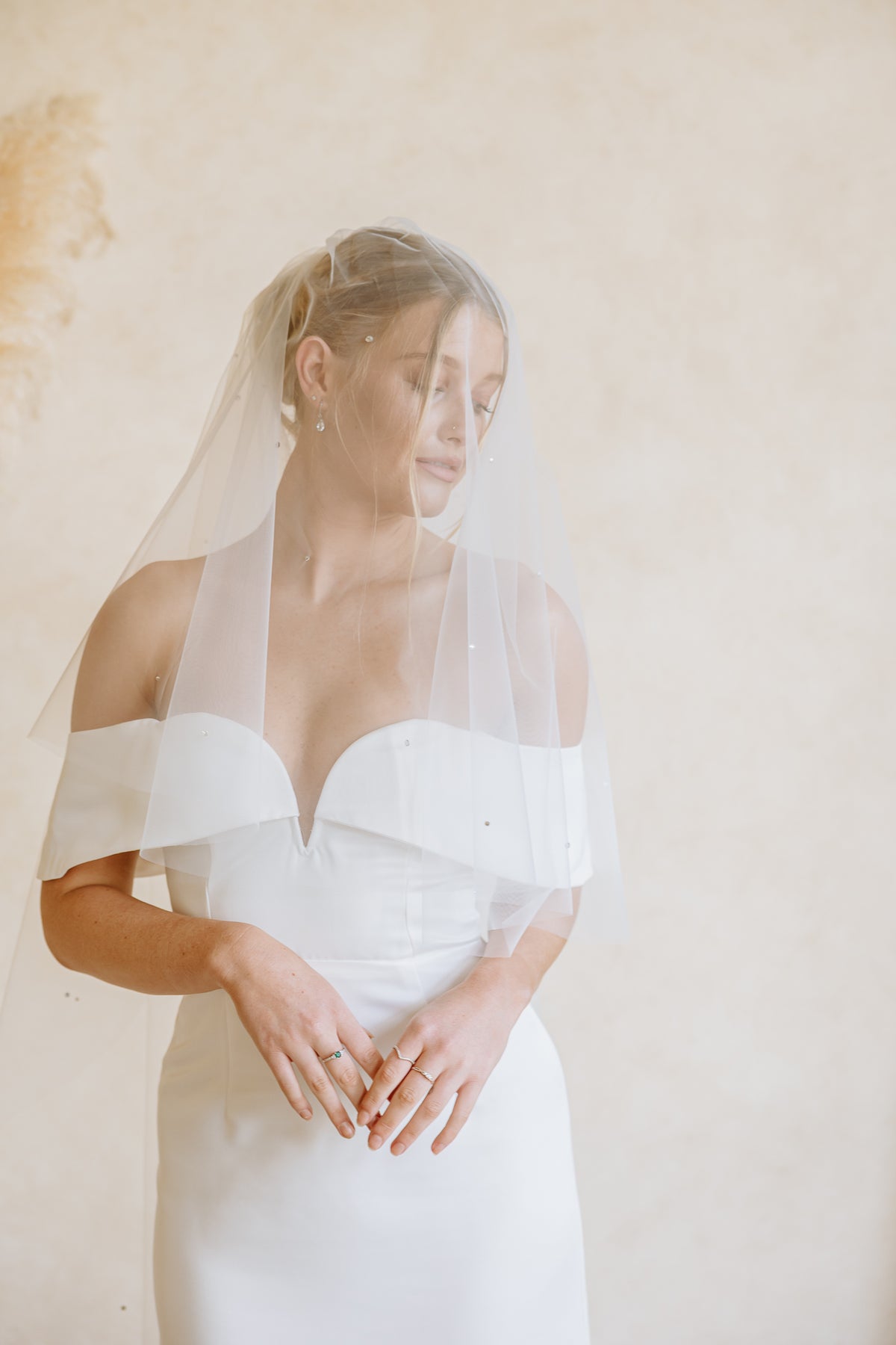 A woman wearing a white off-the-shoulder wedding dress and a sheer white bridal veil, standing against a warm beige textured wall.