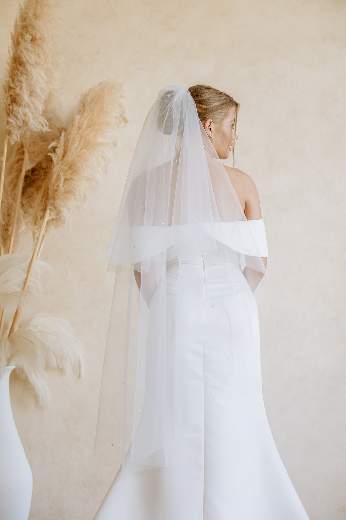 A bride's view from the back, wearing a white off-the-shoulder wedding dress and a long sheer tulle bridal veil, photographed from the back against a warm beige wall with pampas grass decor on the left.