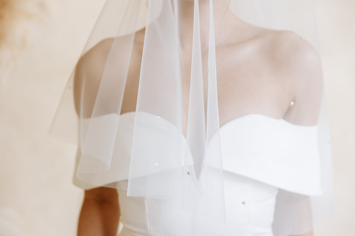 A close-up of a bride wearing a strapless white wedding dress and a long sheer white veil with scattered rhinestones, with the focus on the neckline and fabric details against a soft beige background.