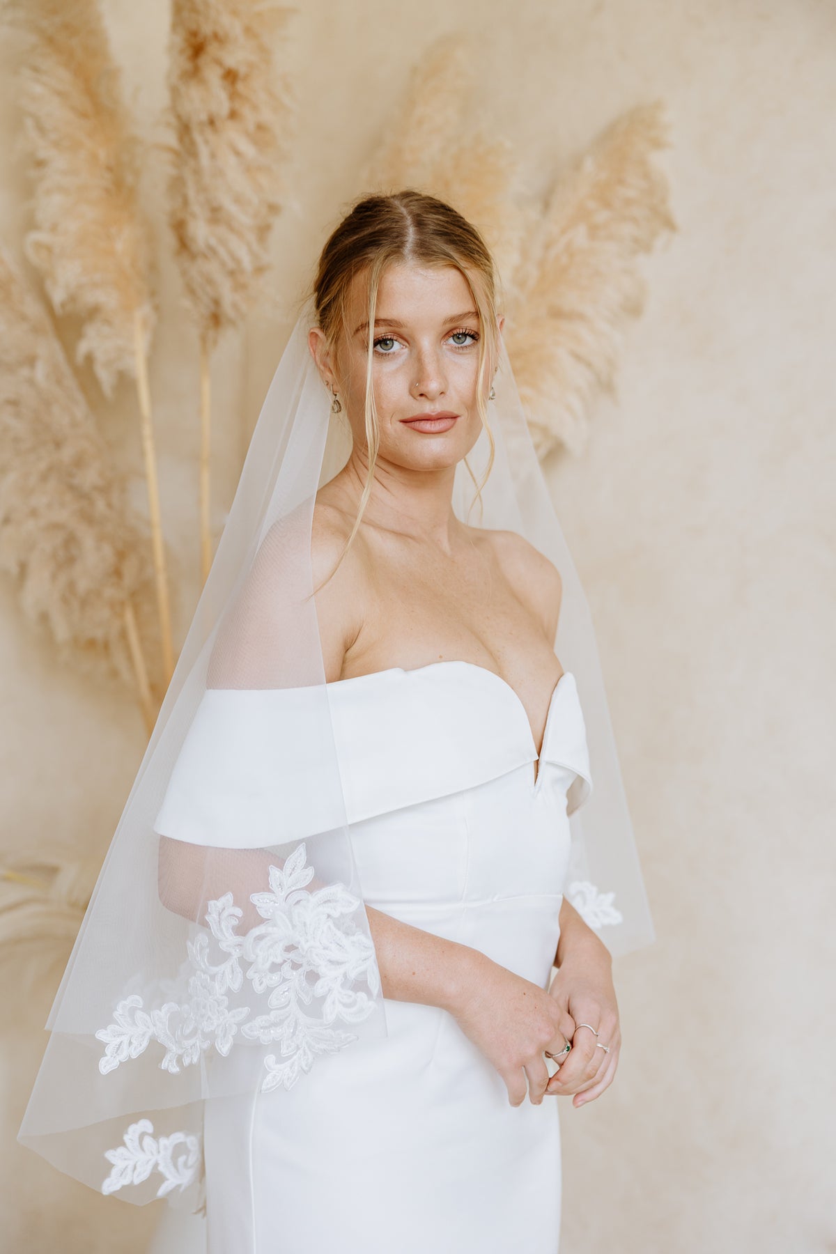 A woman wearing a white off-the-shoulder wedding dress with lace sleeve details and a sheer white veil, standing against a warm beige backdrop with pampas grass.