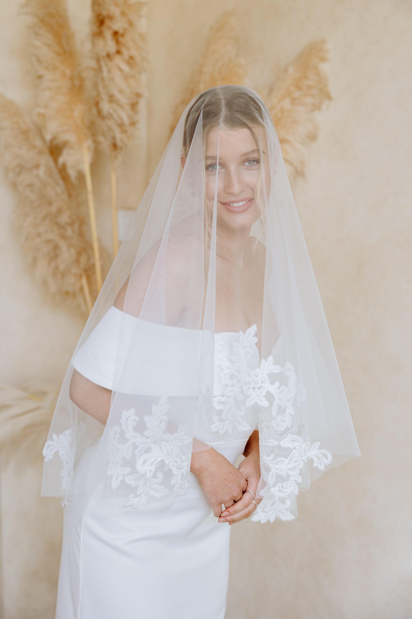 A woman wearing a white off-the-shoulder wedding dress with lace sleeve details and a sheer white veil, standing against a warm beige backdrop with pampas grass.