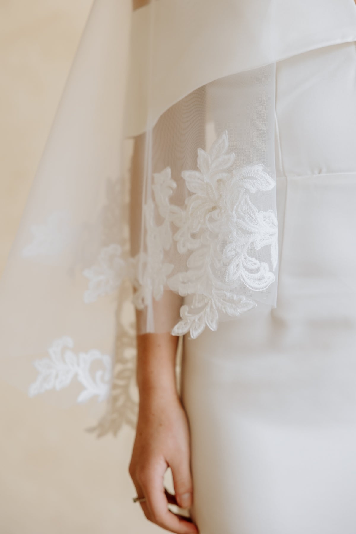 Close-up of a white bridal gown with sheer sleeves featuring intricate white floral lace applique, worn by a bride whose hand is visible holding the lace.