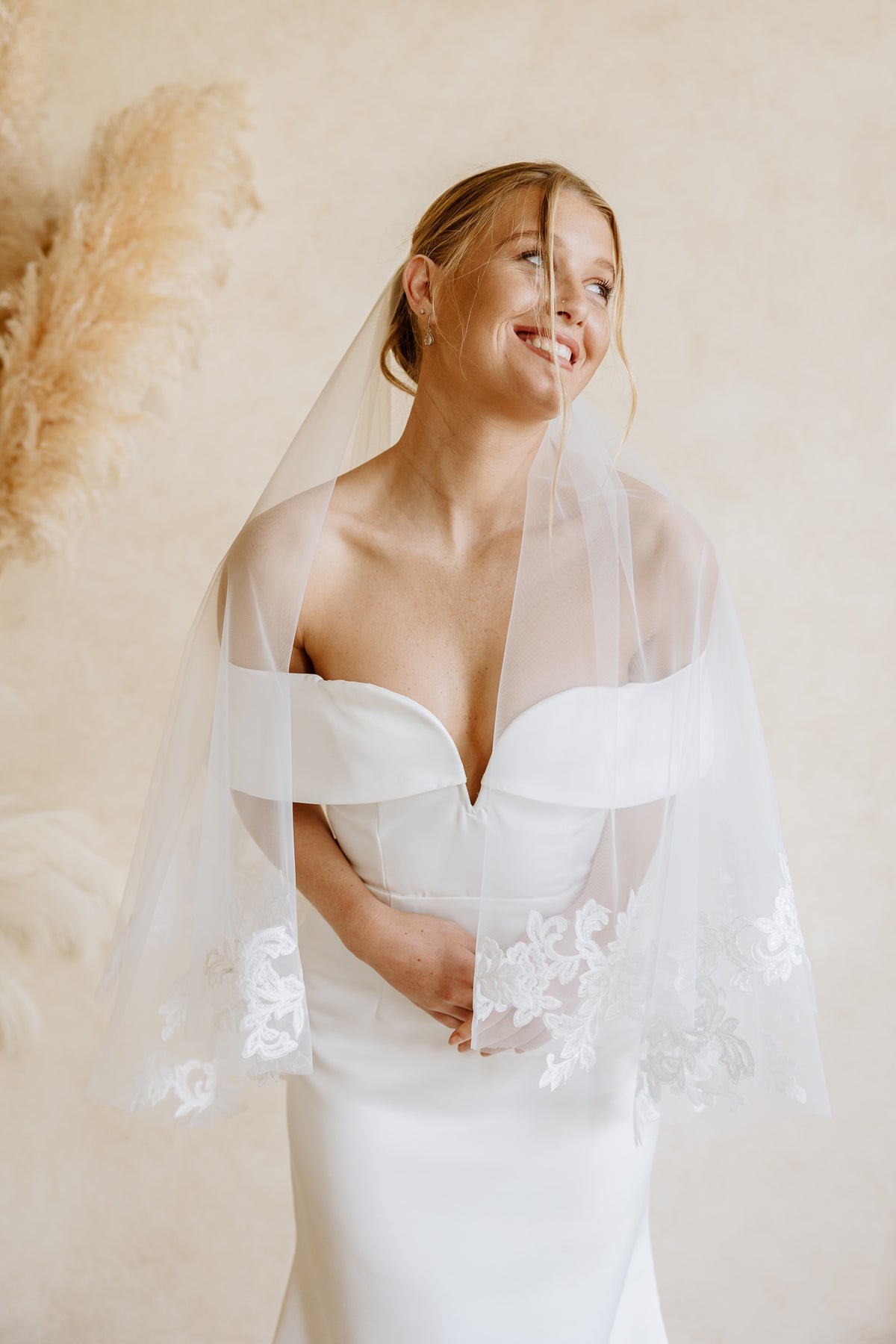 A smiling bride wearing a white strapless wedding dress and a sheer white veil with floral lace trim, posed against a warm beige wall with pampas grass decor.