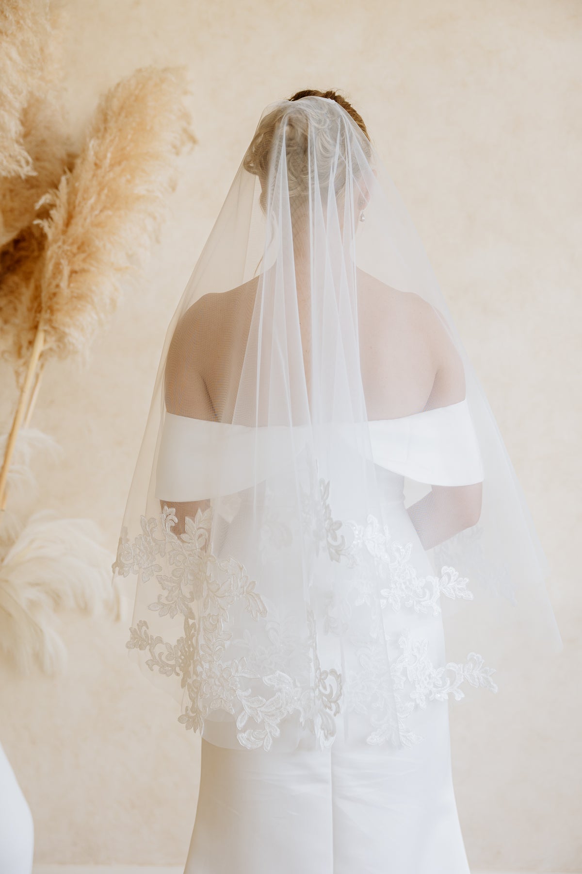 A bride viewed from behind wearing a long, sheer white tulle bridal veil over an off-the-shoulder white wedding dress with lace detailing, set against a warm beige wall with pampas grass decor.